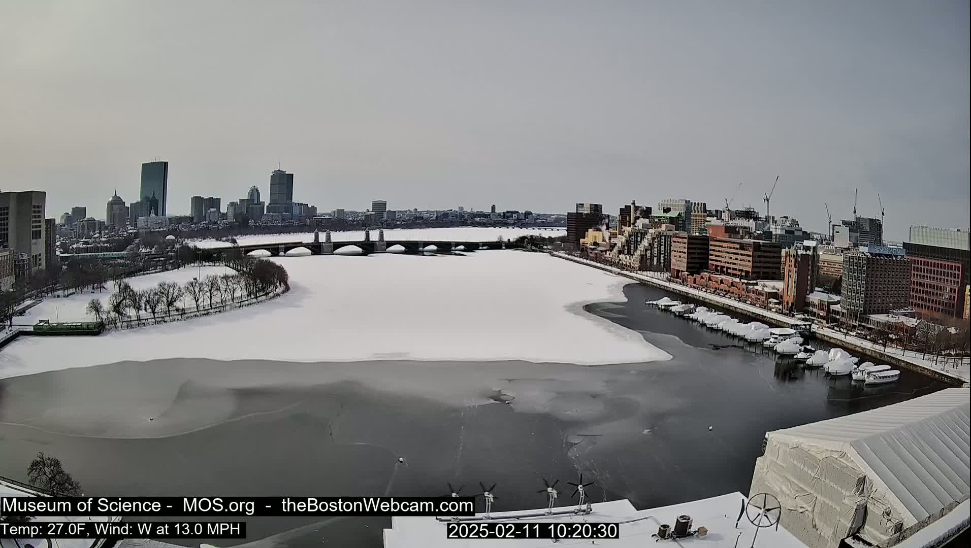 A snow-covered river with a bridge and a city skyline in the background, under a cloudy sky.