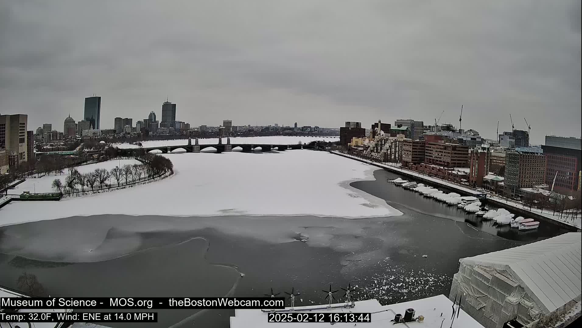 A partially frozen river flows past a city skyline under an overcast sky.