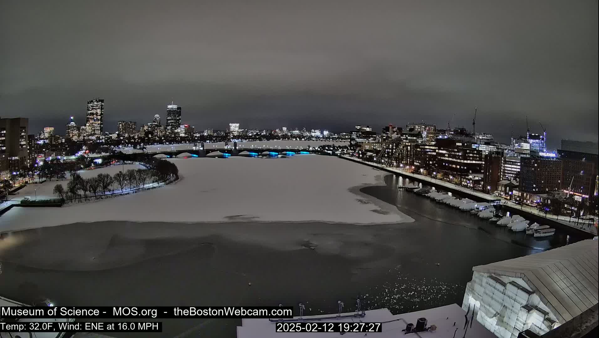 A nighttime cityscape with a partially frozen river, under a cloudy sky, showing city lights reflecting on the water.