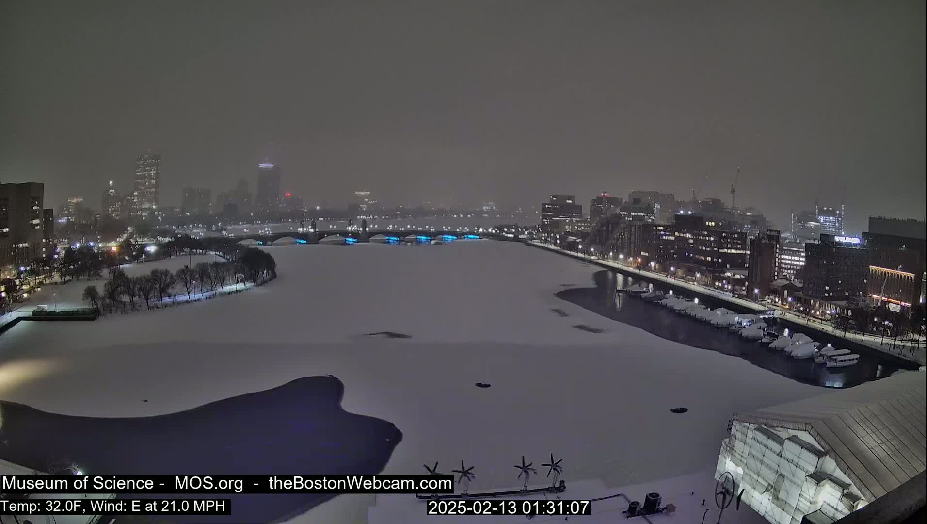 A snow-covered city waterfront at night, with a partially frozen river, is seen under a hazy, dark sky with an east wind at 21 mph and a temperature of 32 degrees Fahrenheit.