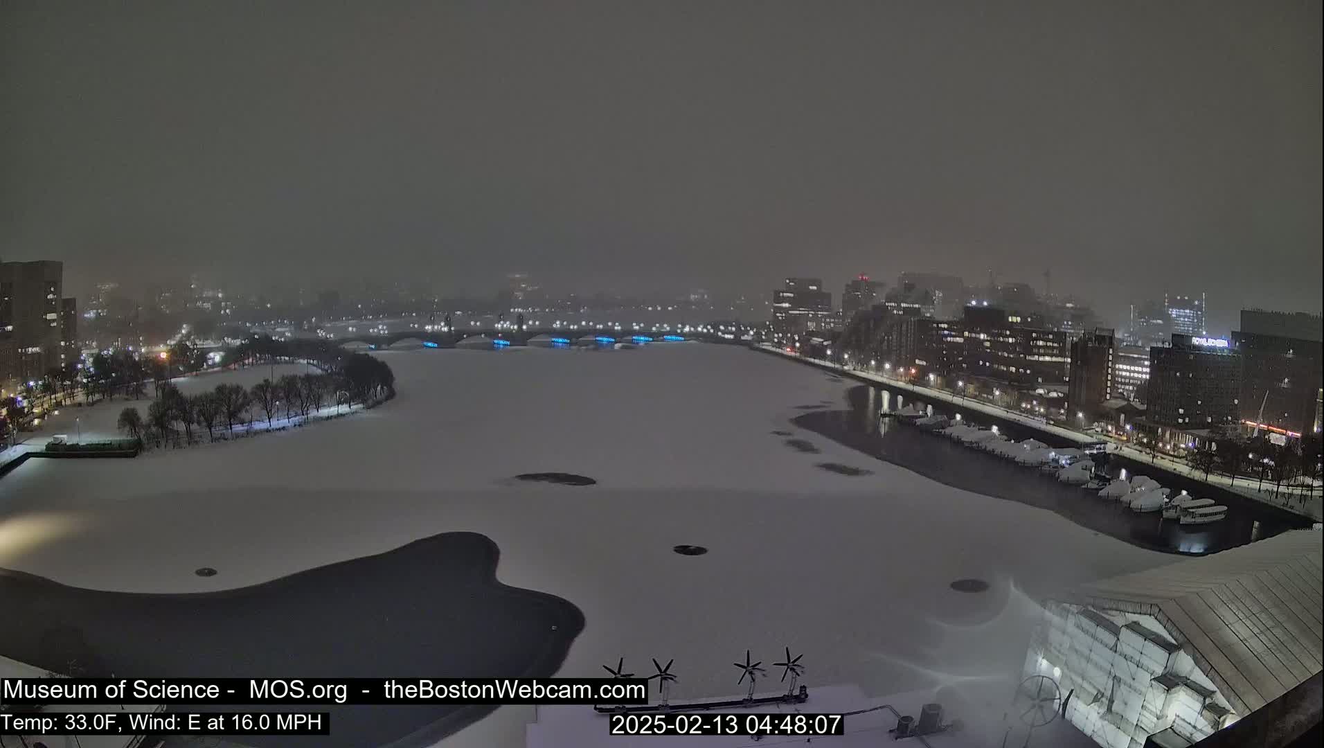 A snow-covered cityscape at night, with a partially frozen body of water, under a dark, snowy sky.