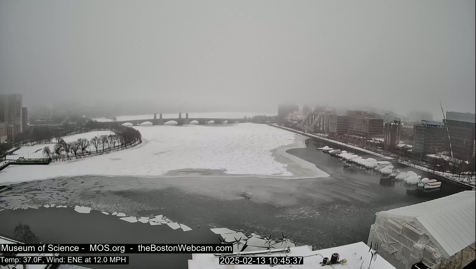 A snow-covered cityscape is seen across a partially frozen river with broken ice floes, under a hazy, gray sky.