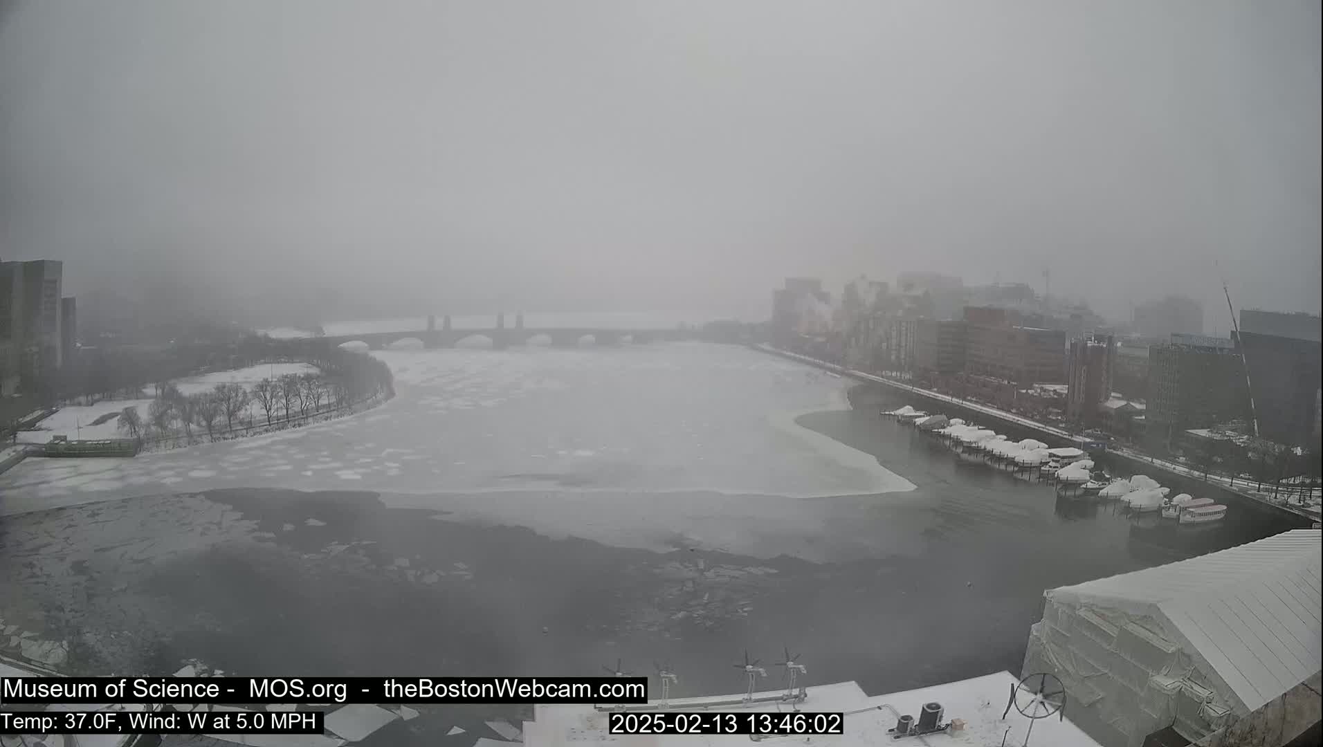 A snow-covered river with partially frozen water flows past a bridge and city buildings under a hazy, overcast sky.
