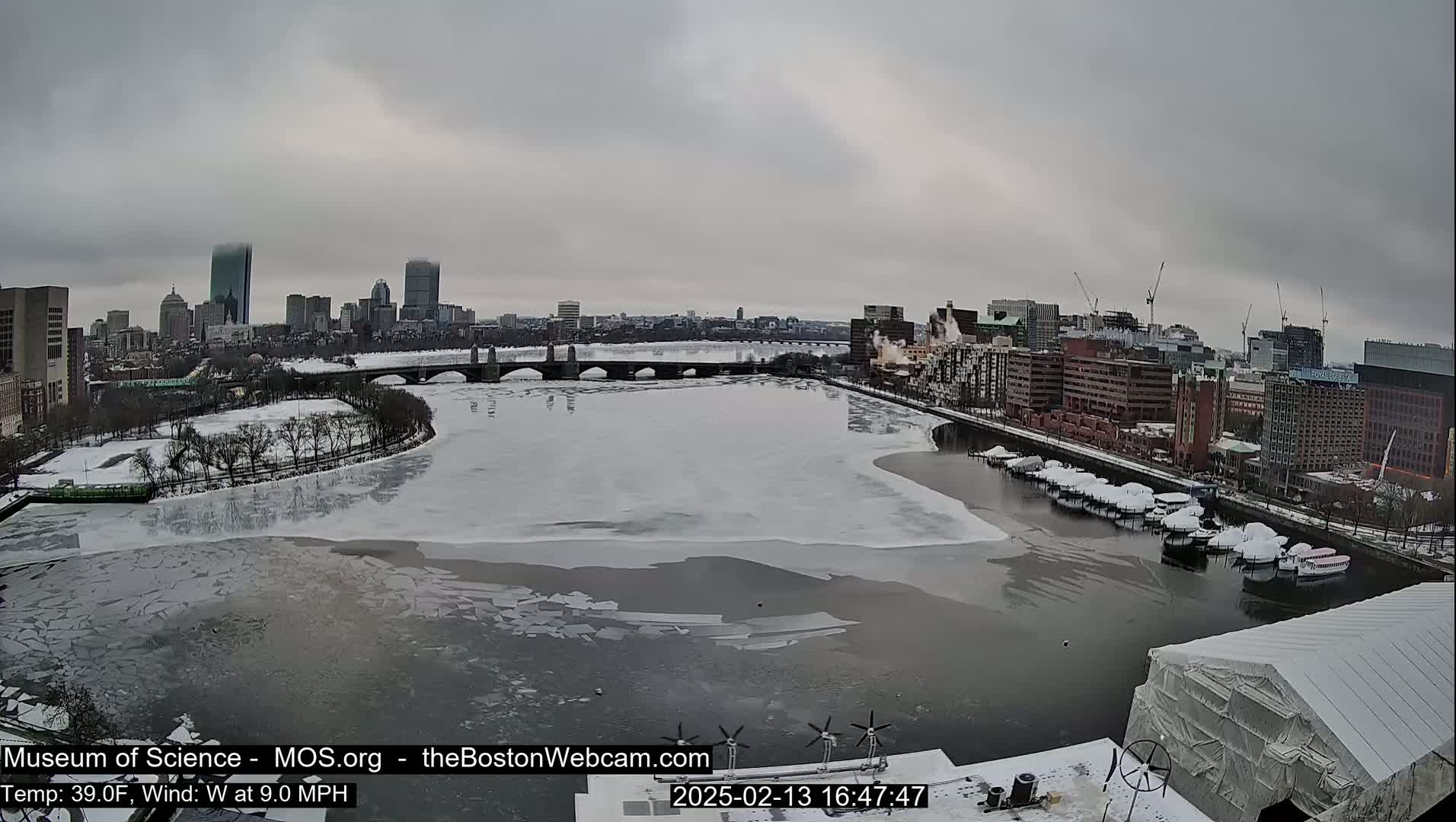 A partly frozen river flows through a snowy city under an overcast sky.