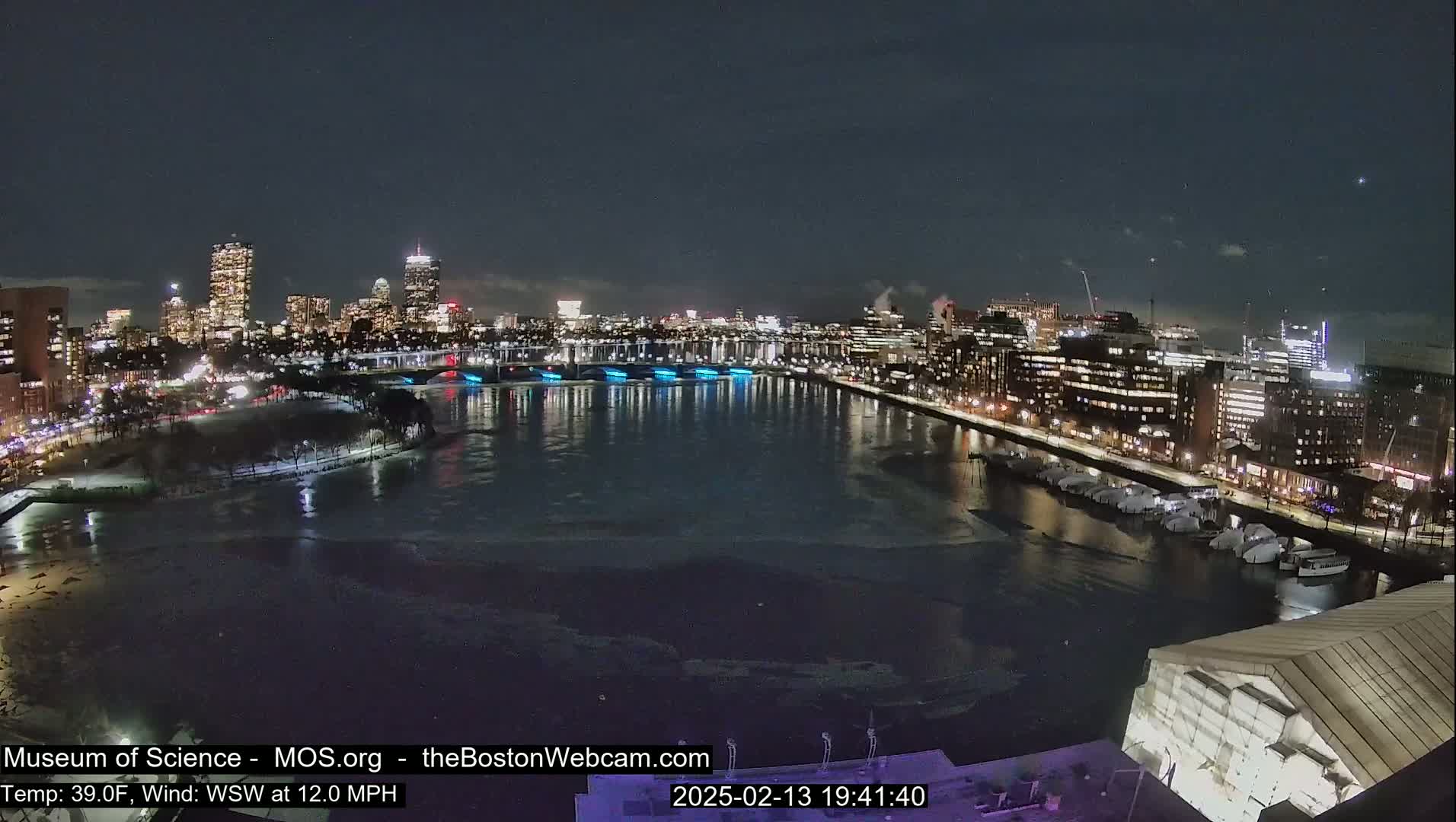 A nighttime view of a city skyline across a partially ice-covered river, with a wind of 12 mph from the west-southwest and a temperature of 39°F.