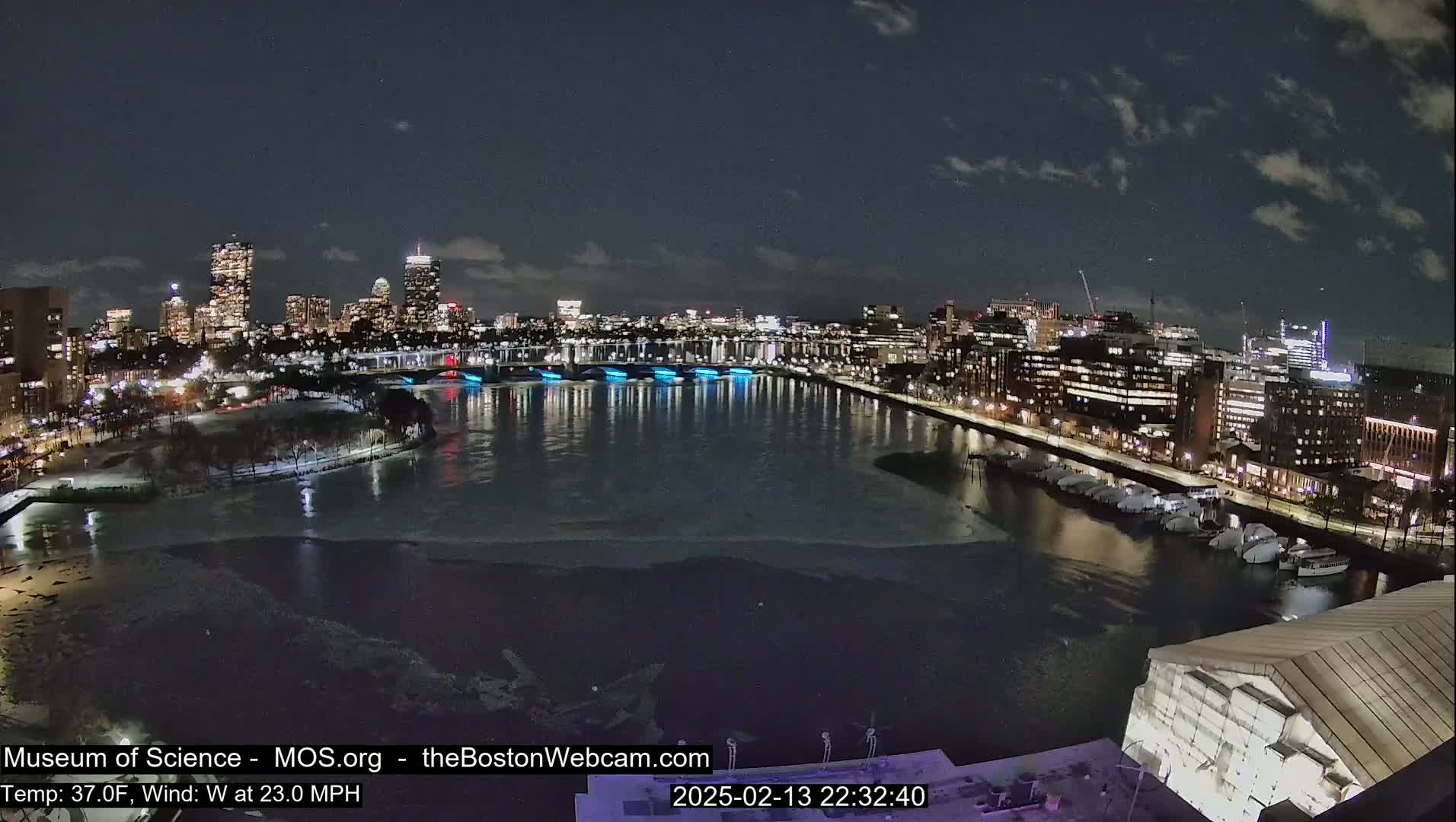 A nighttime cityscape with a partially frozen river, illuminated bridges, and a waterfront lined with buildings under a partly cloudy sky.