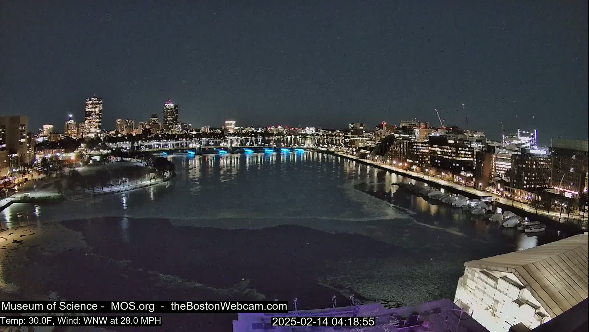 A nighttime cityscape with a partially frozen river reflecting city lights, under a clear, dark sky with a strong wind.