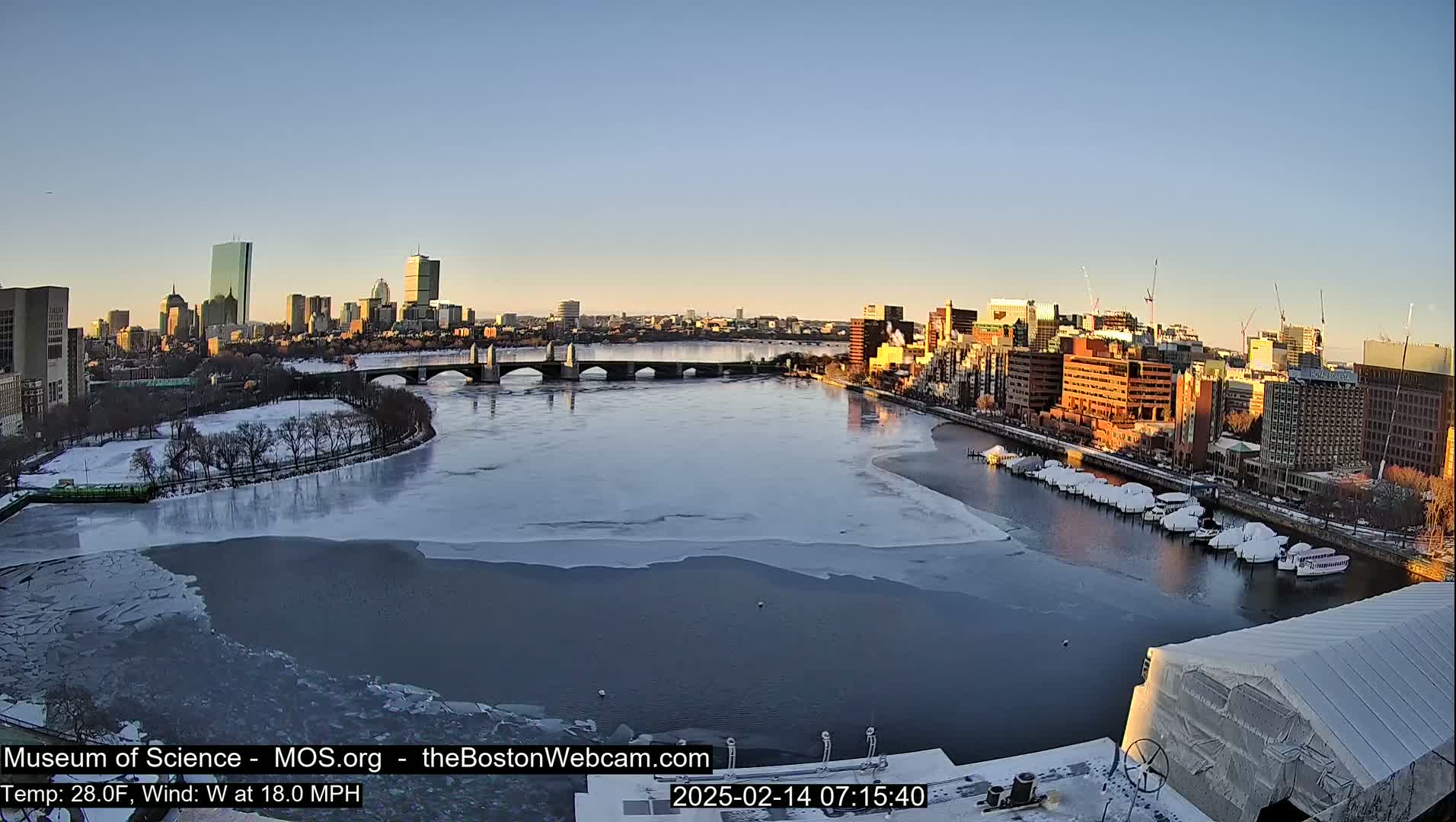 A partially frozen river flows past a city skyline at sunrise, with a bridge spanning the river and boats docked along the shore.