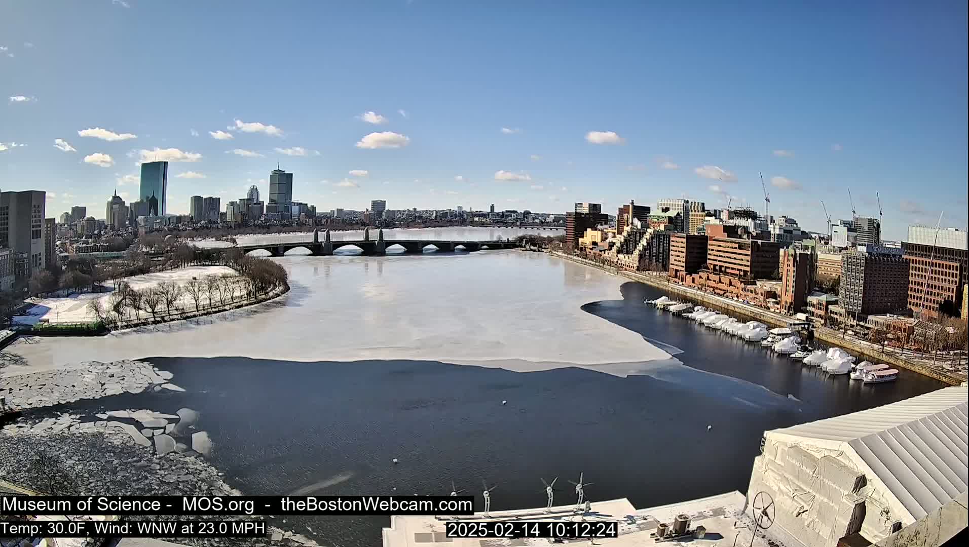 A partially frozen river flows through a city, with a bridge and numerous buildings visible under a sunny sky.