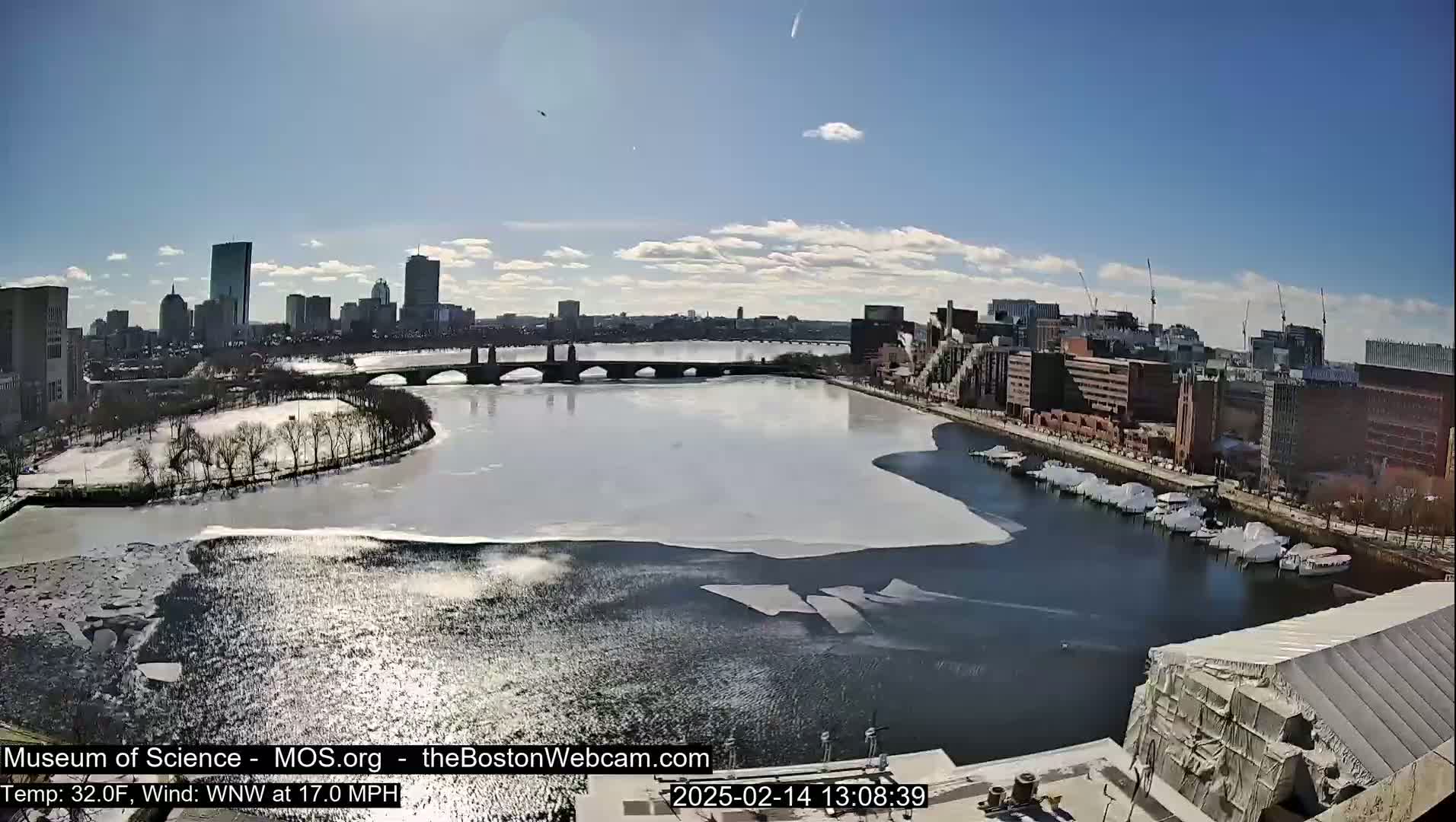 A partly sunny day shows a city skyline across a partially frozen river with a bridge and docked boats, under a clear blue sky.