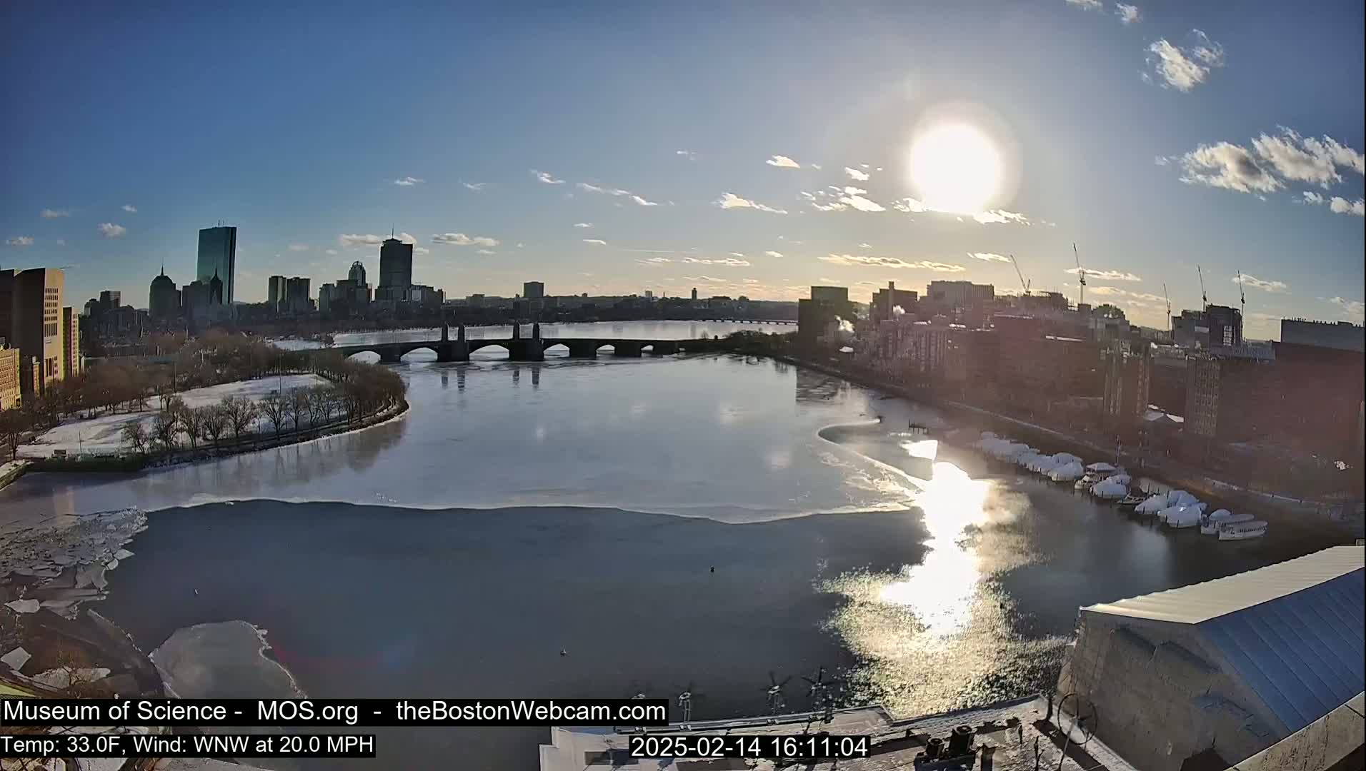 A partially frozen river flows beneath a bridge, with a city skyline visible in the background under a bright, sunny sky.
