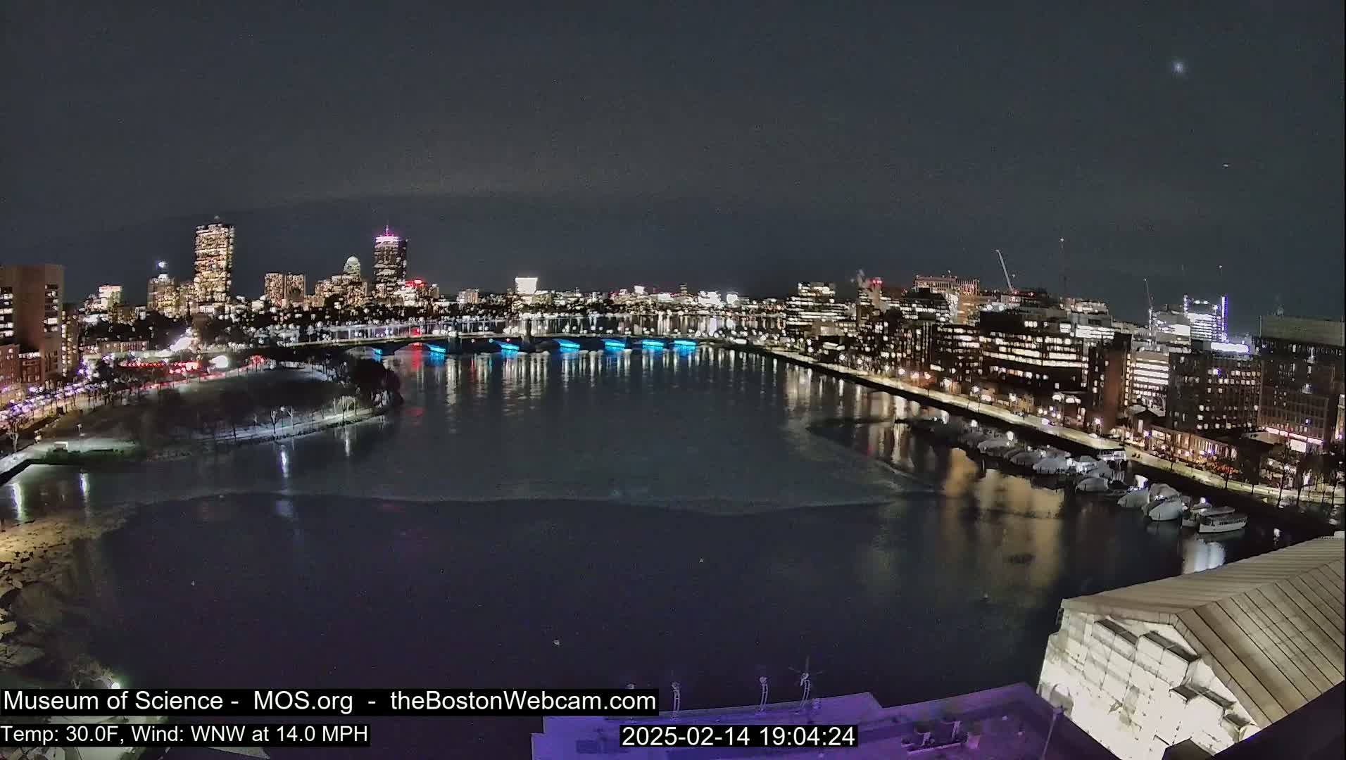 A nighttime view of a city skyline across a partially frozen river, with a 30°F temperature and a 14 mph wind from the west-northwest.