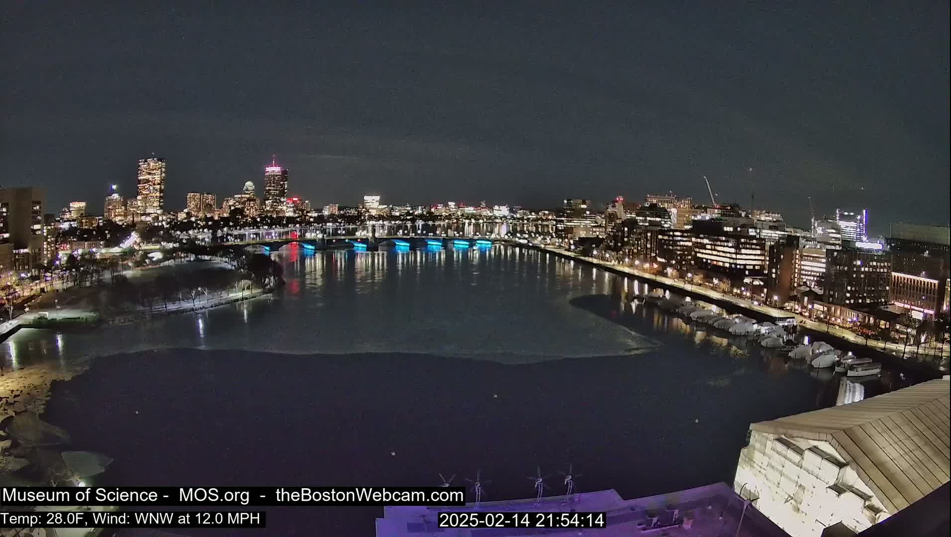 A nighttime view of a city skyline across a partially frozen river, with a brisk wind.