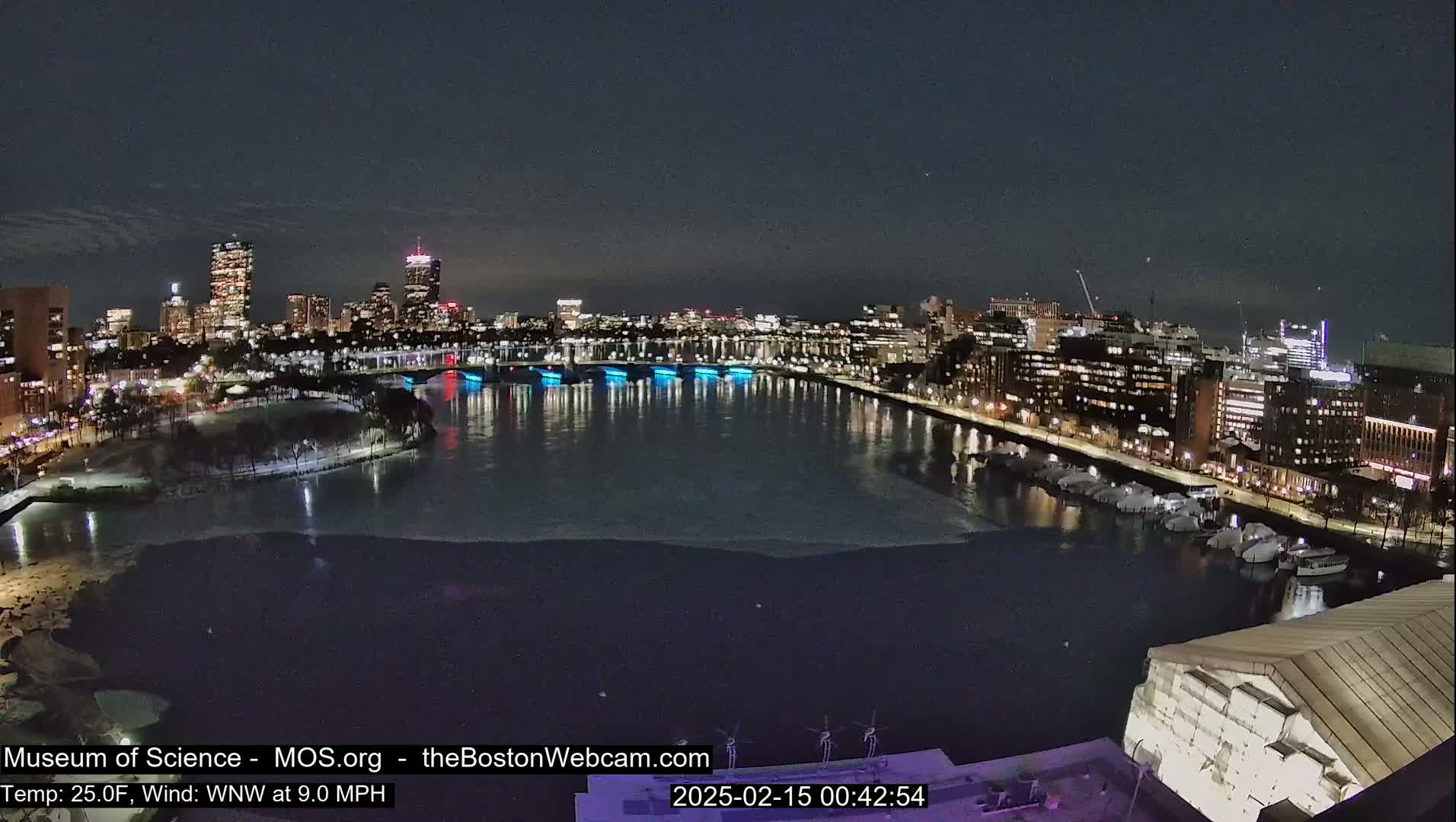 A nighttime view of a city skyline across a partially frozen river, with a 25°F temperature and a 9 mph wind from the west-northwest.