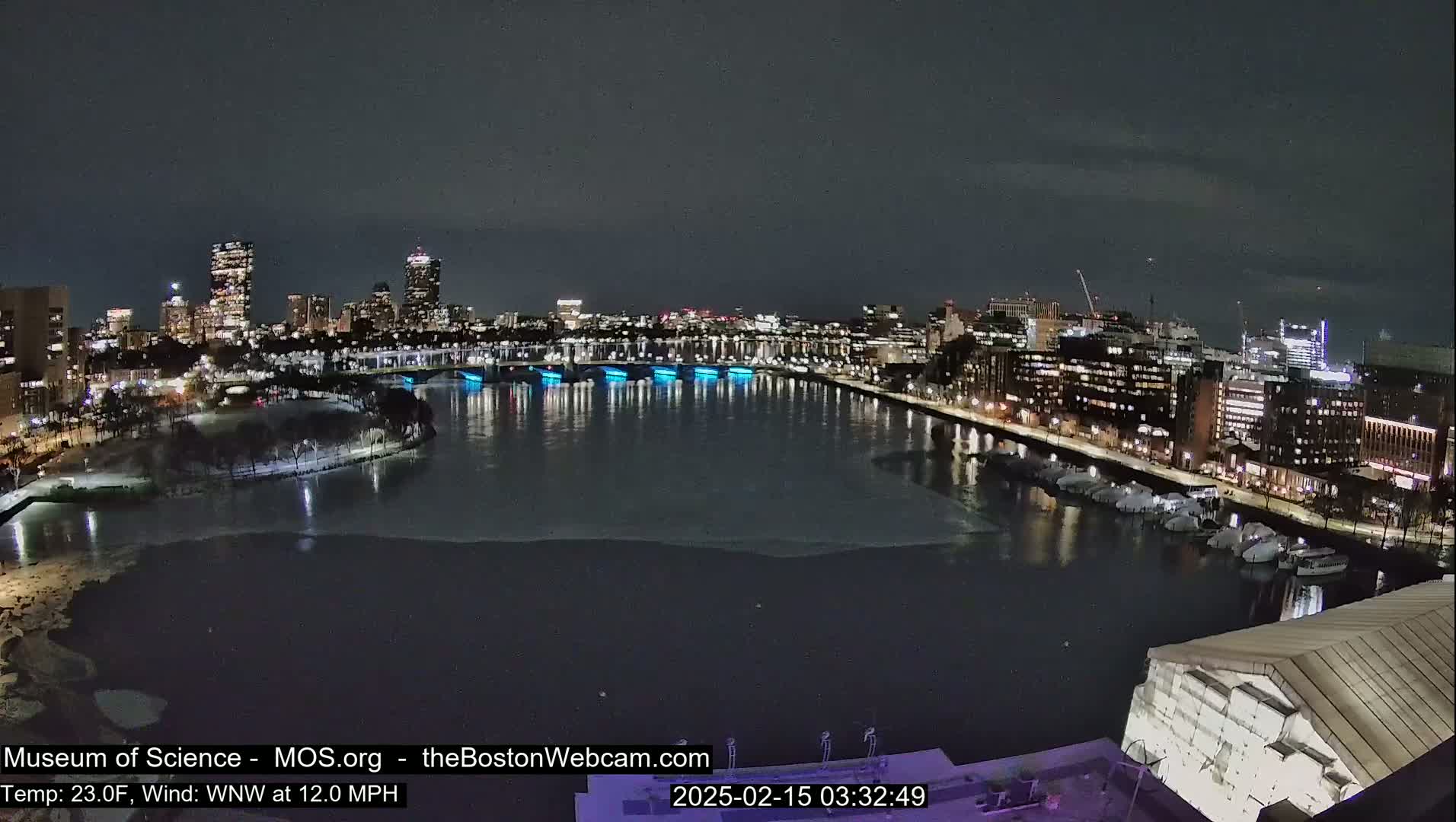 A nighttime view of a city skyline across a partially frozen river, under a dark sky.