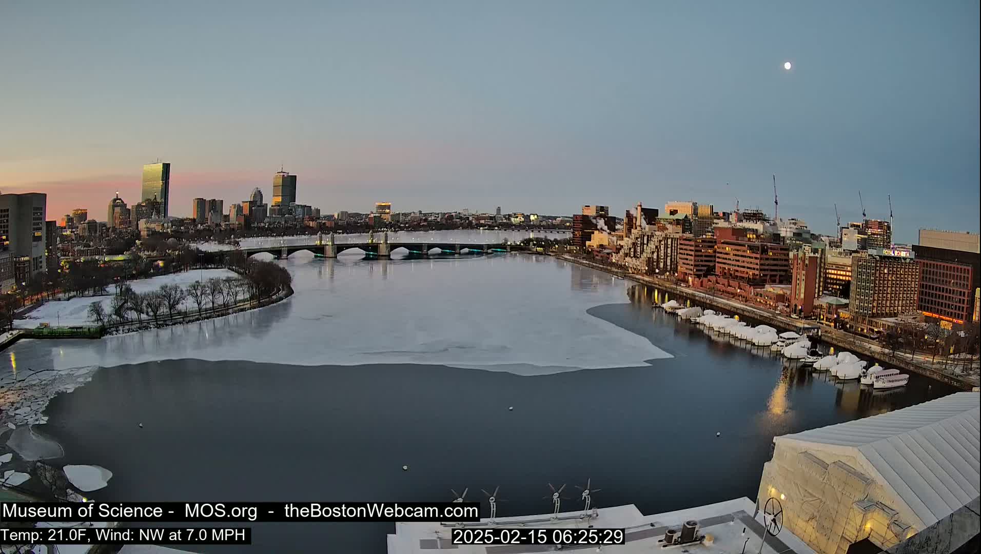 A partially frozen river flows through a city at dawn under a clear sky with a visible moon, showcasing a bridge and a cityscape with several tall buildings.