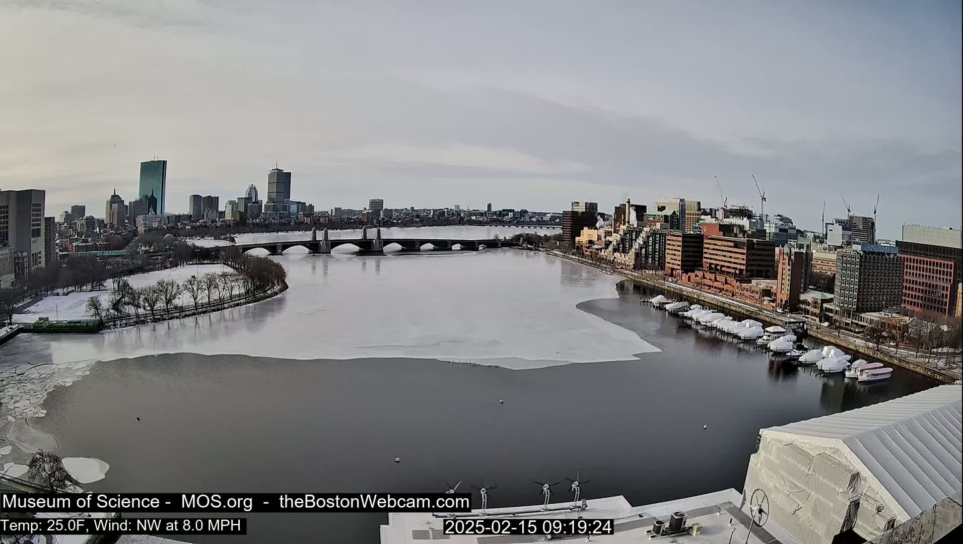 A partially frozen river flows past a city skyline with a bridge, boats covered in white, and snow-covered trees under a mostly clear sky.
