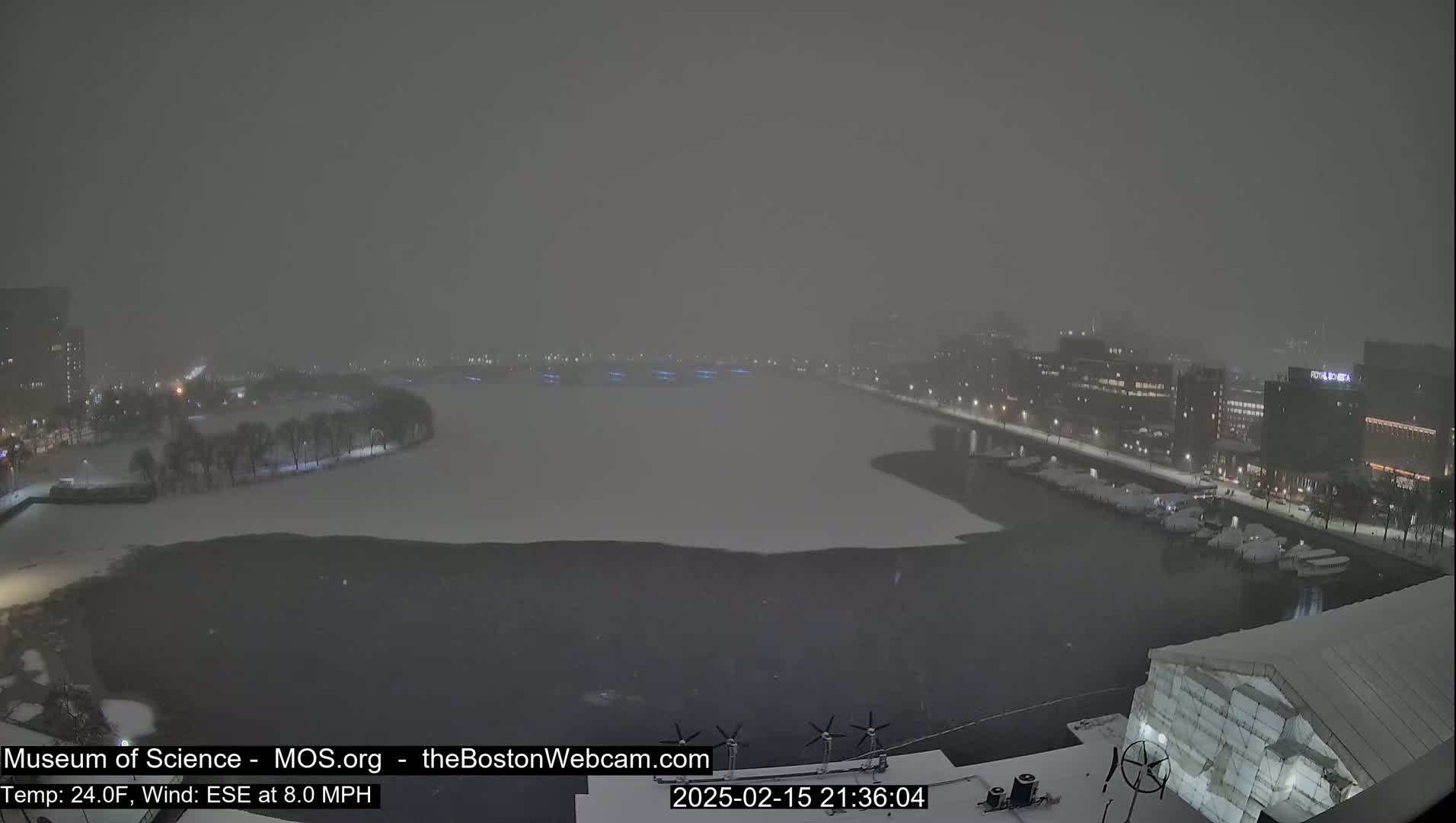 A snow-covered waterfront scene at night, with buildings and boats visible through falling snow.