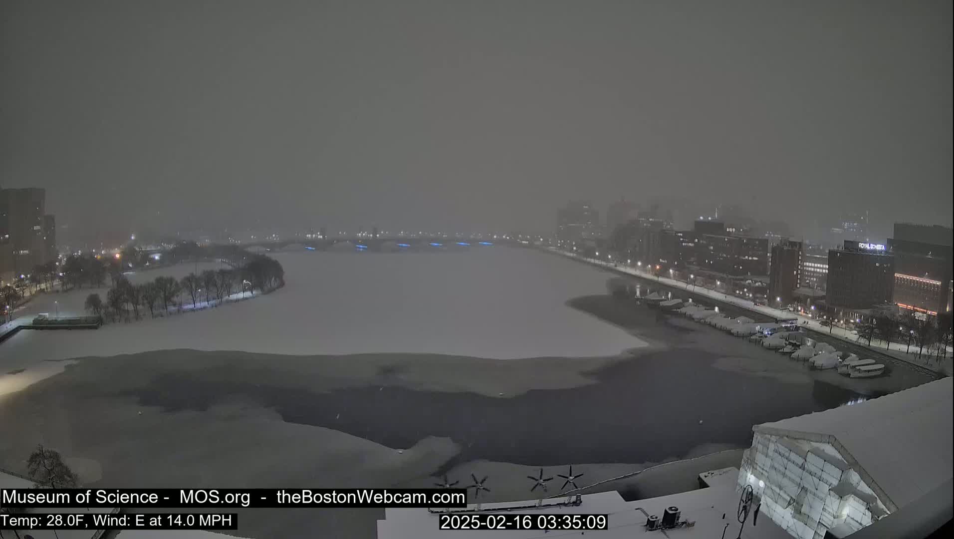 A snowstorm blankets a city waterfront at night, with a partially frozen river visible between snow-covered parkland and buildings.