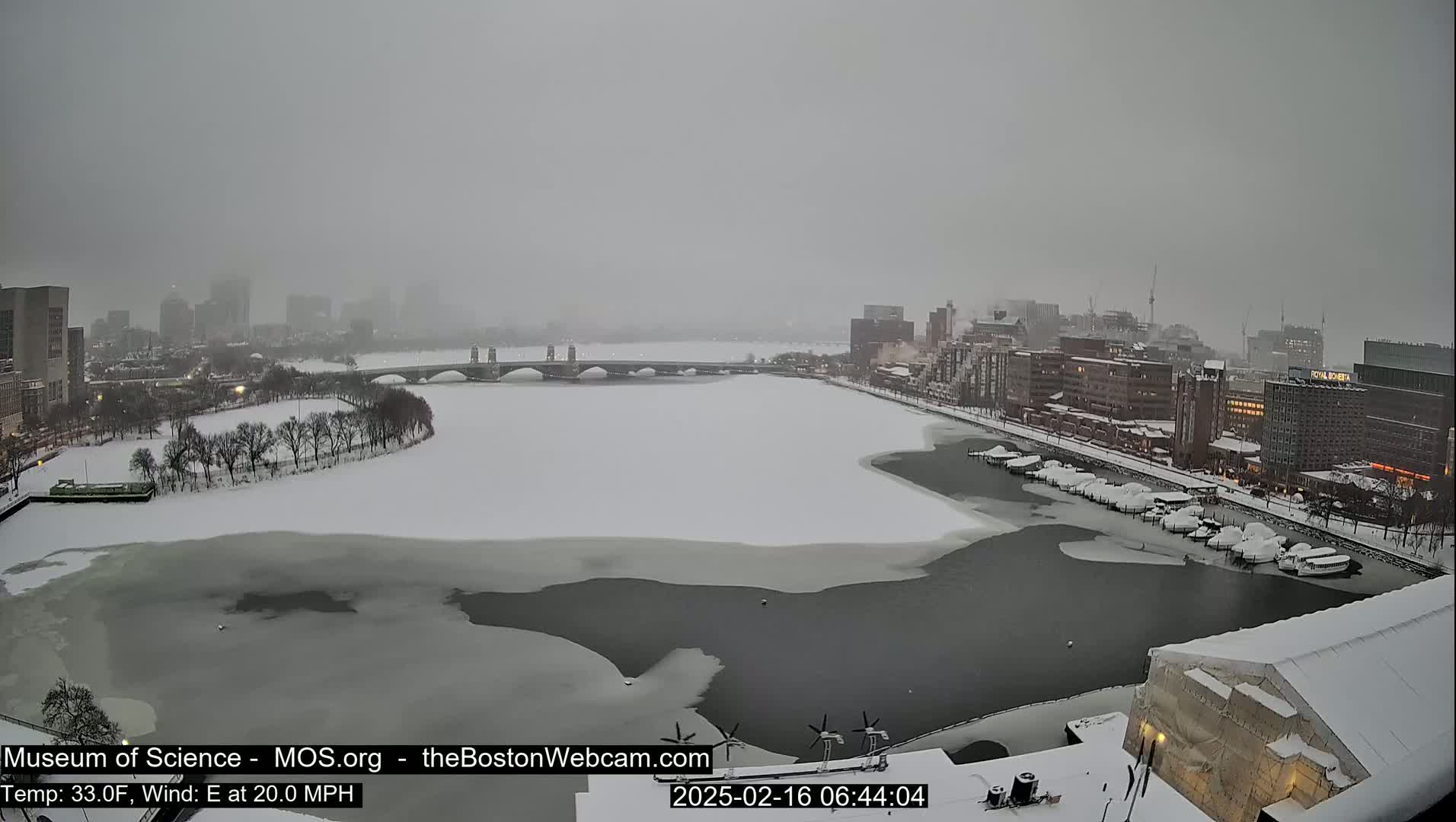 A snow-covered cityscape with a partially frozen river, a bridge, and boats docked along the shore during a snowy day with an easterly wind.