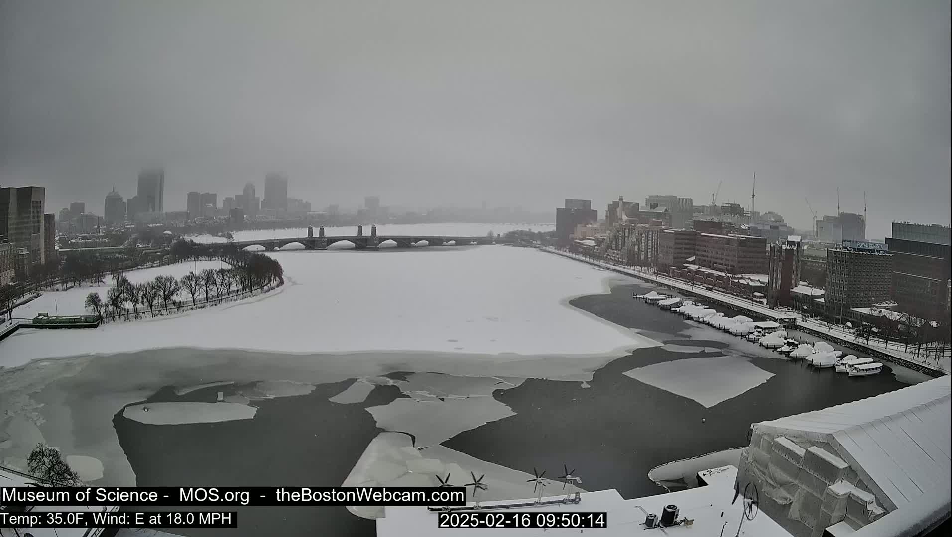 A snow-covered city waterfront with a partially frozen river, a bridge, and numerous buildings under an overcast sky.