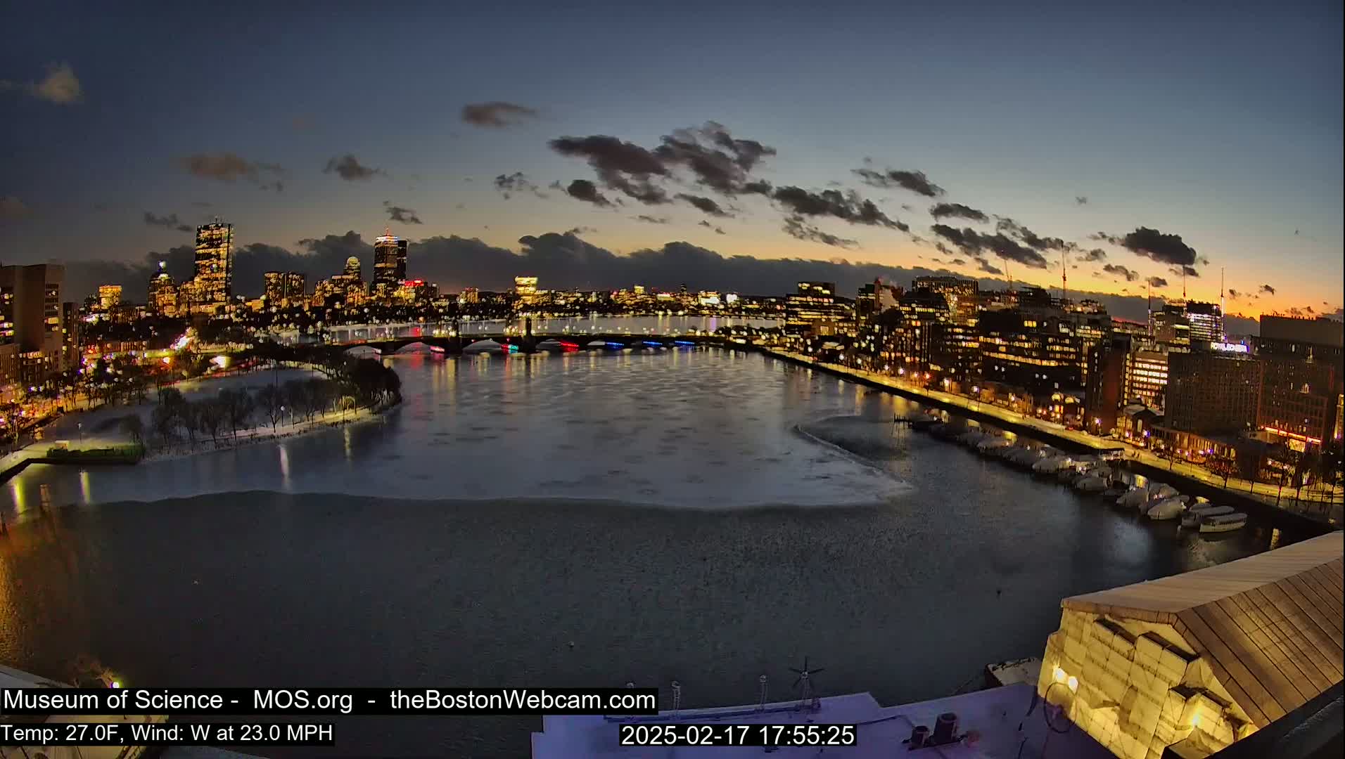 A nighttime city skyline with a partially frozen river, under a partly cloudy sky.