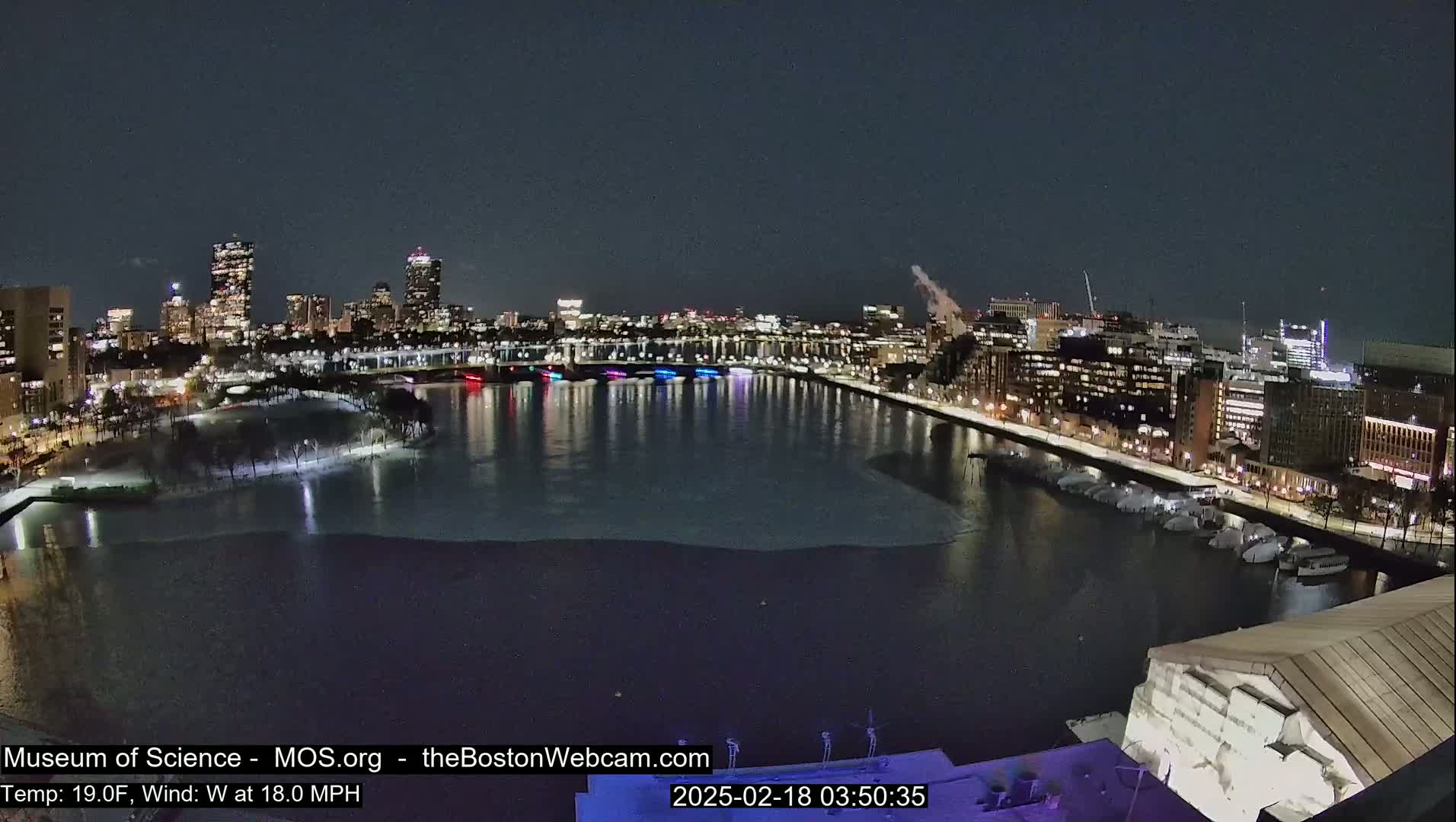 A nighttime view of a city skyline across a partially frozen river, with a cold wind blowing at 18 mph.