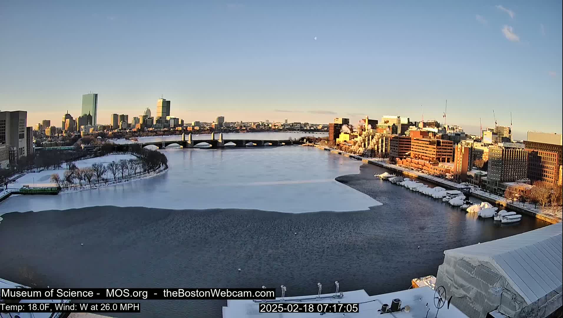 A partially frozen river flows through a city under a clear, bright sky, with a bridge spanning the waterway and buildings covered in snow.
