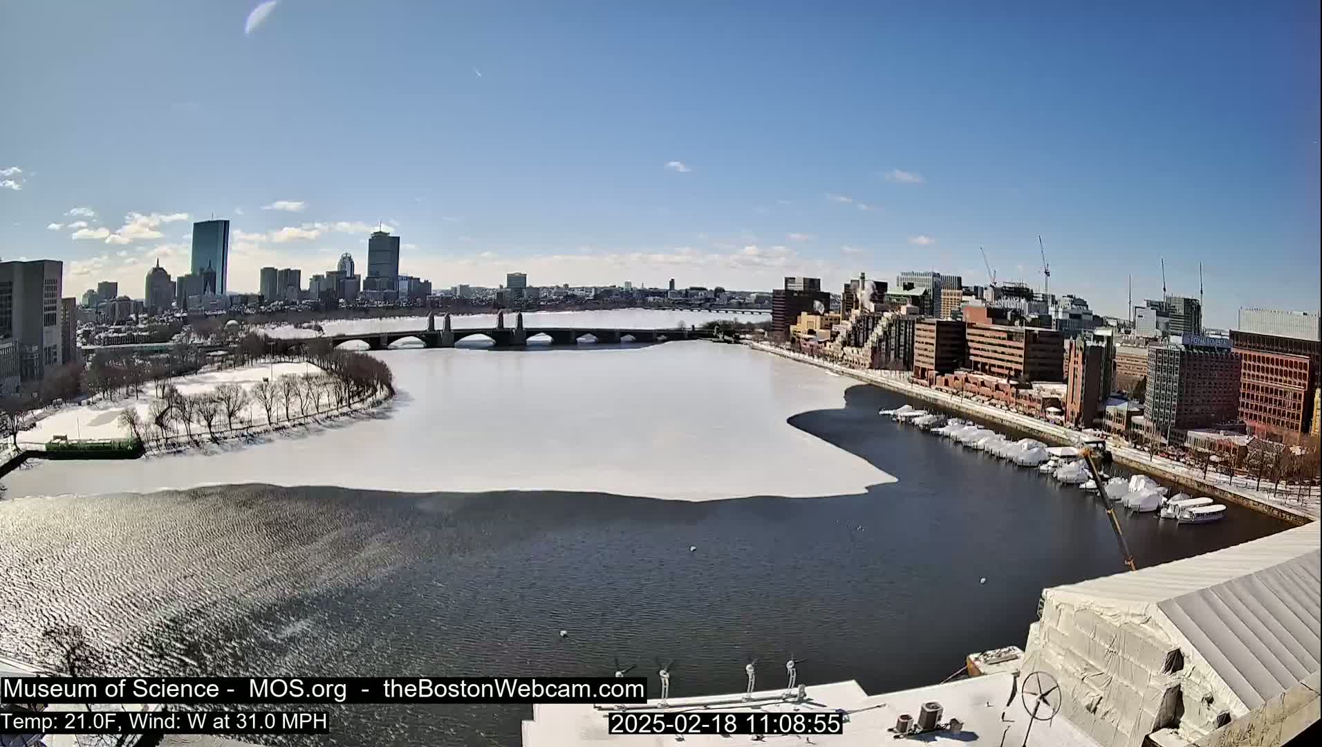 A partially frozen river flows through a city, under a clear blue sky, with a bridge spanning the river and snow covering the ground and some of the water.