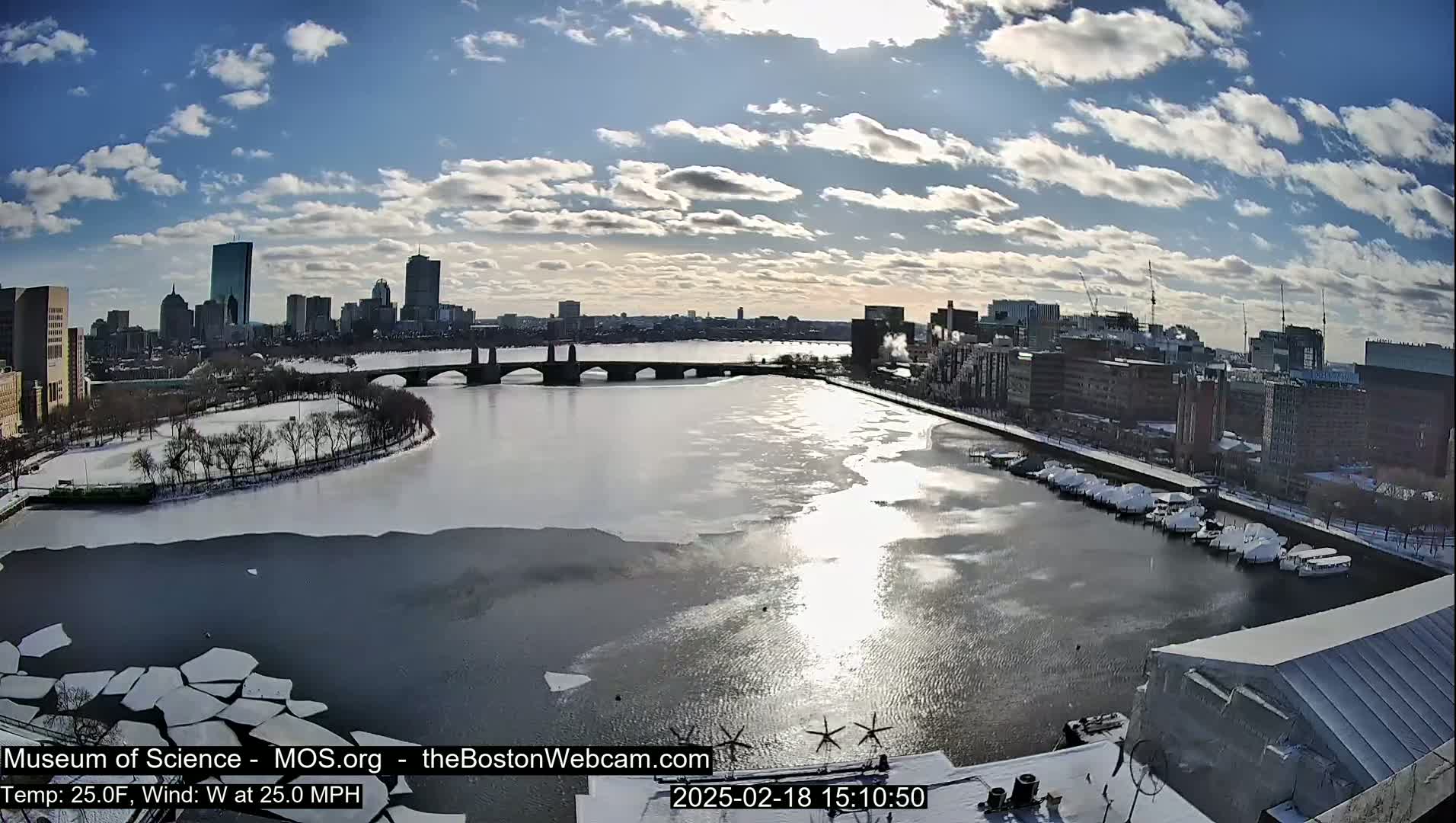 A partially frozen river flows past a city skyline under a partly sunny sky, with snow visible on the ground and boats covered in snow at a marina.