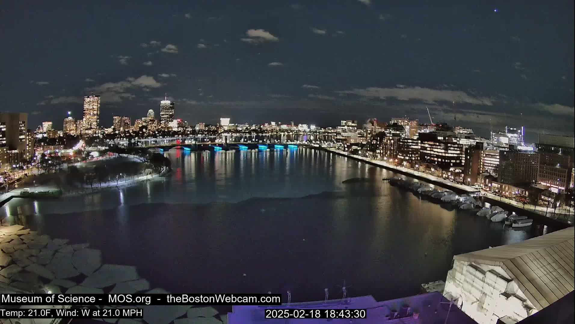 A nighttime view of a city skyline across a partially ice-covered river with a 21°F temperature and a 21 mph westerly wind.
