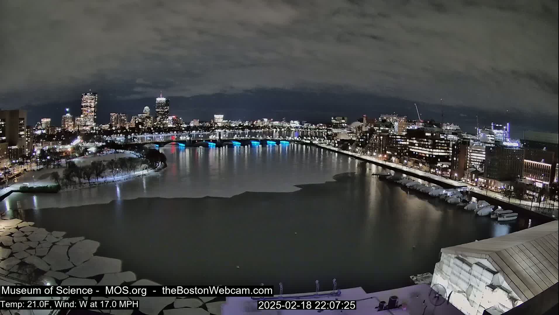A nighttime cityscape with a partially frozen river, showing a bridge with blue lights, under a cloudy sky, and a  wind of 17 mph.