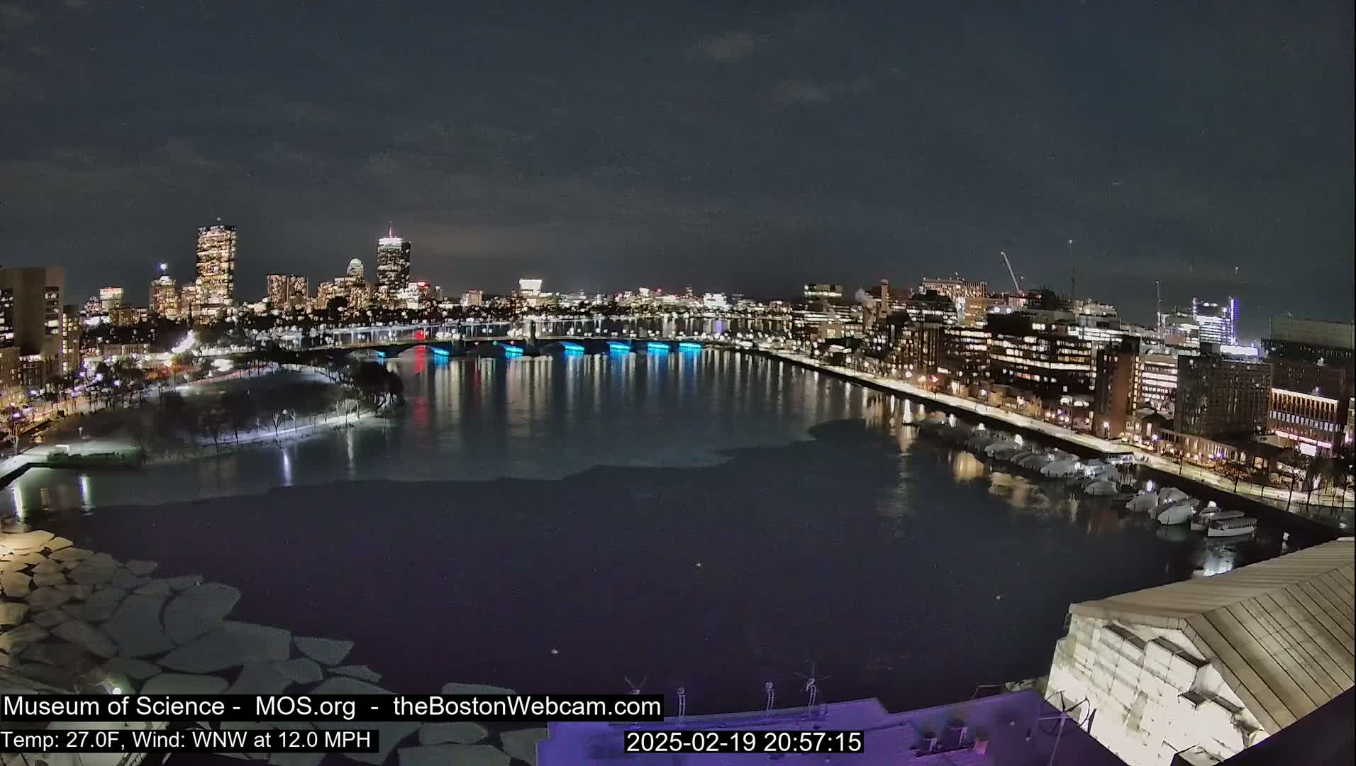 A nighttime cityscape with a river partially frozen over, illuminated buildings, and boats docked along the shore.