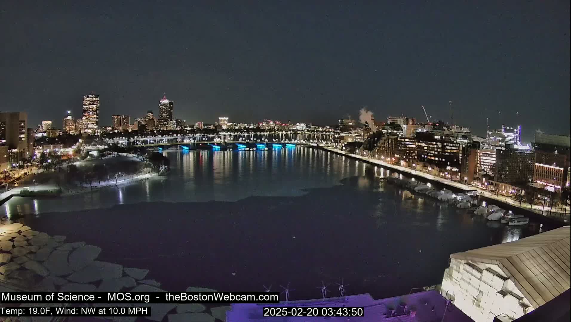 A nighttime cityscape across a partially frozen river, with city lights reflecting on the water under a clear, dark sky.