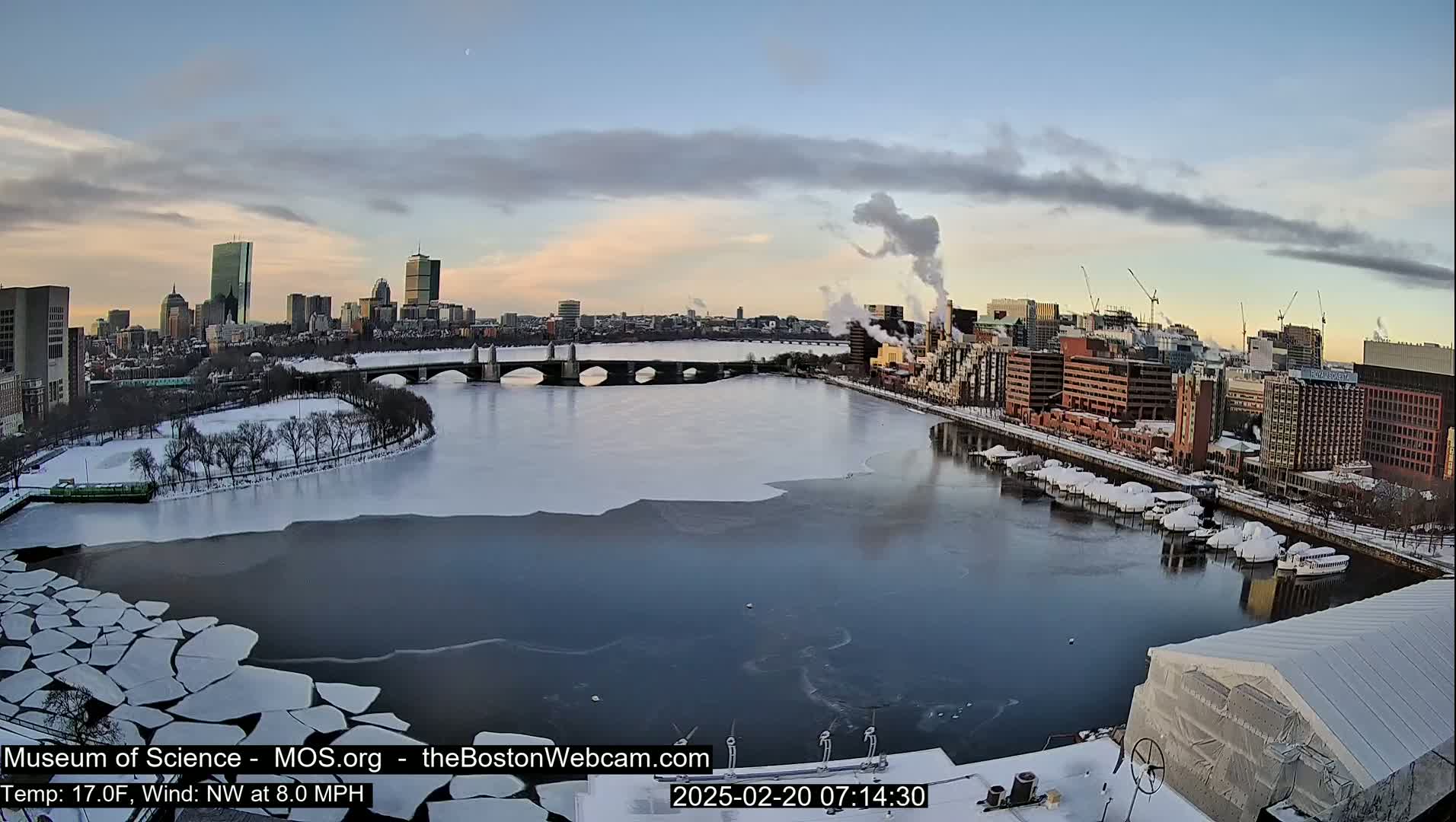 A partly frozen river flows past a snow-covered city, under a partly cloudy sky at sunrise.