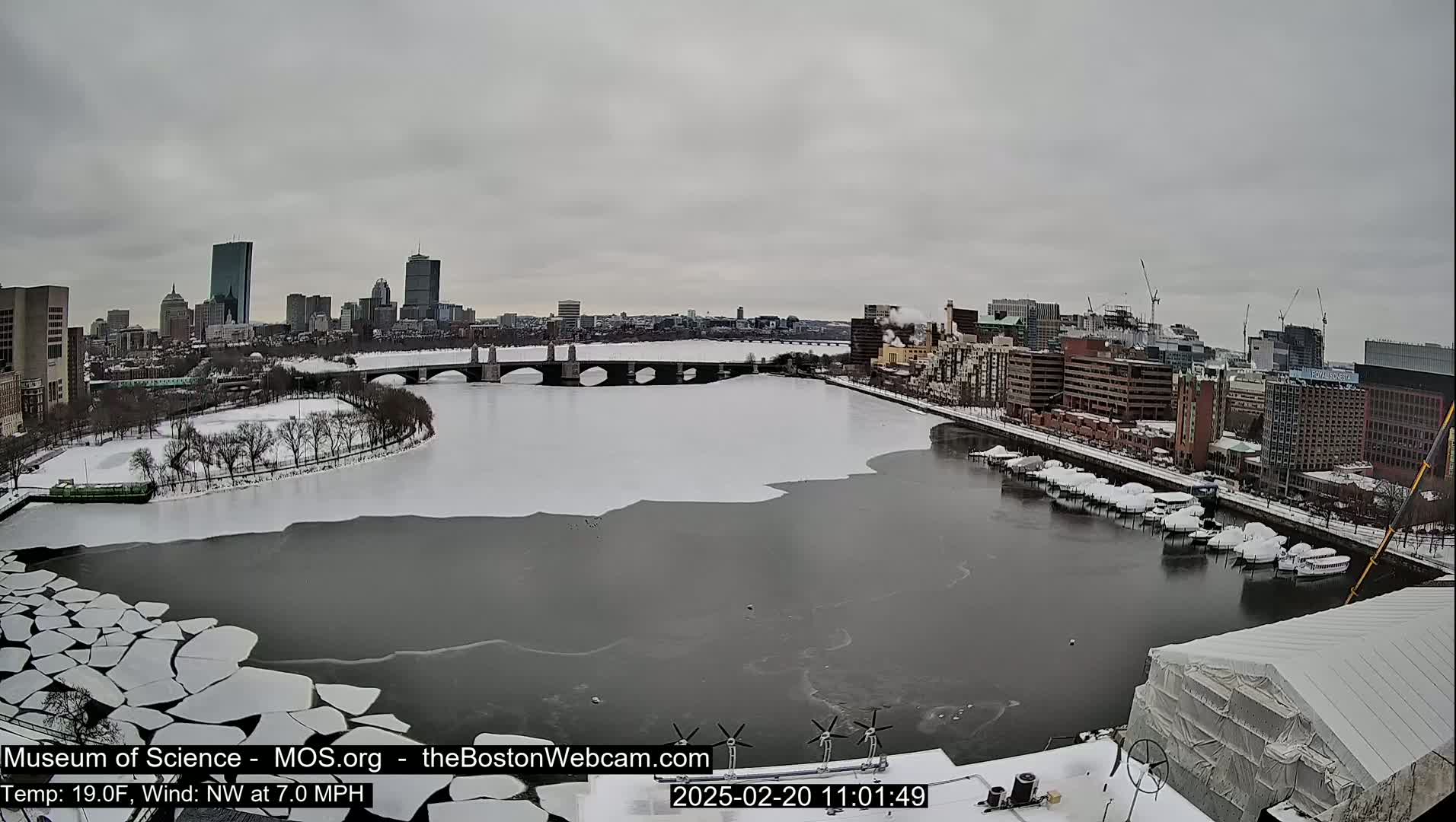 A partially frozen river flows past a snow-covered city under a gray sky.