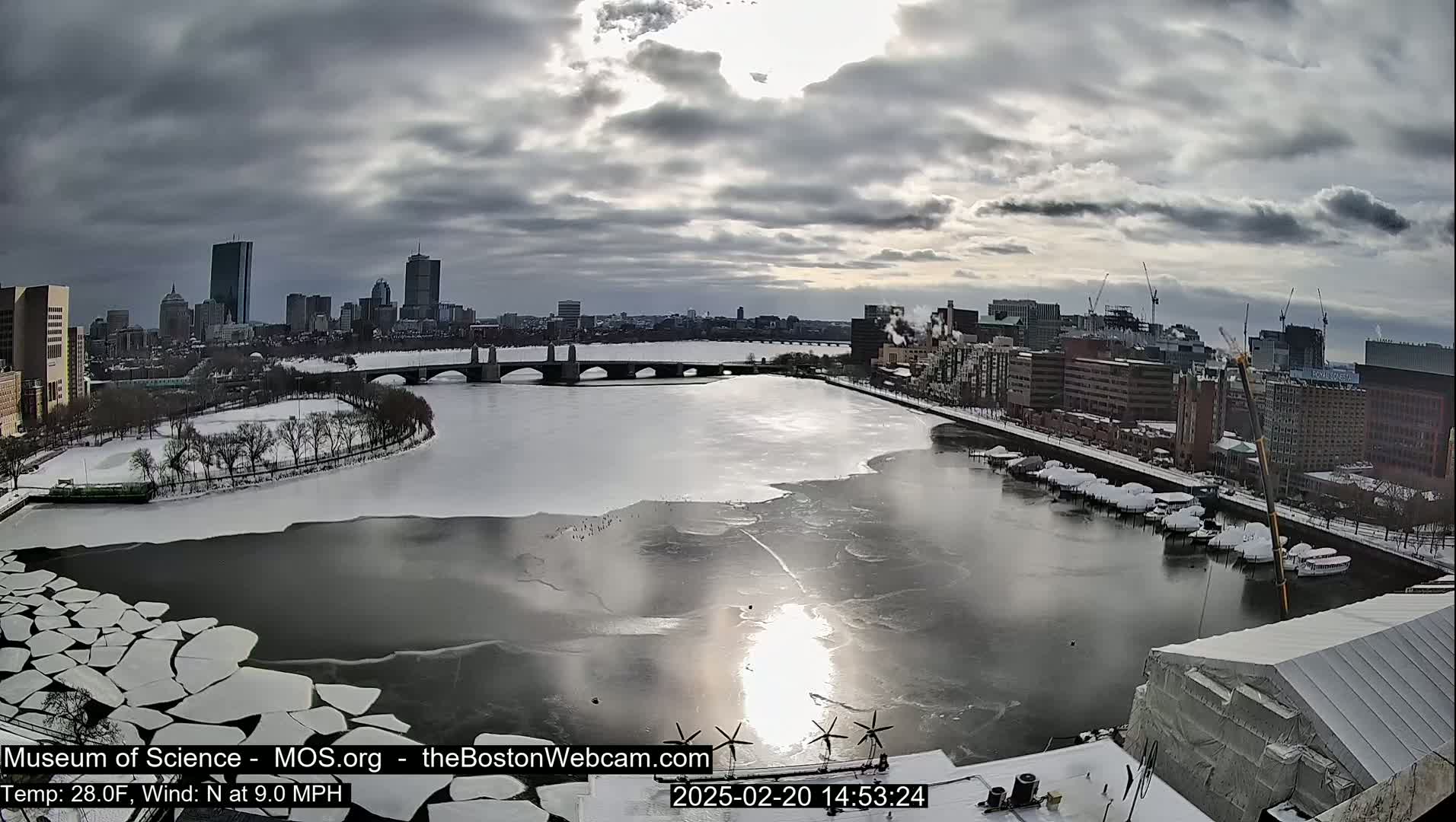 A partially frozen river flows through a snow-covered city under a cloudy sky.