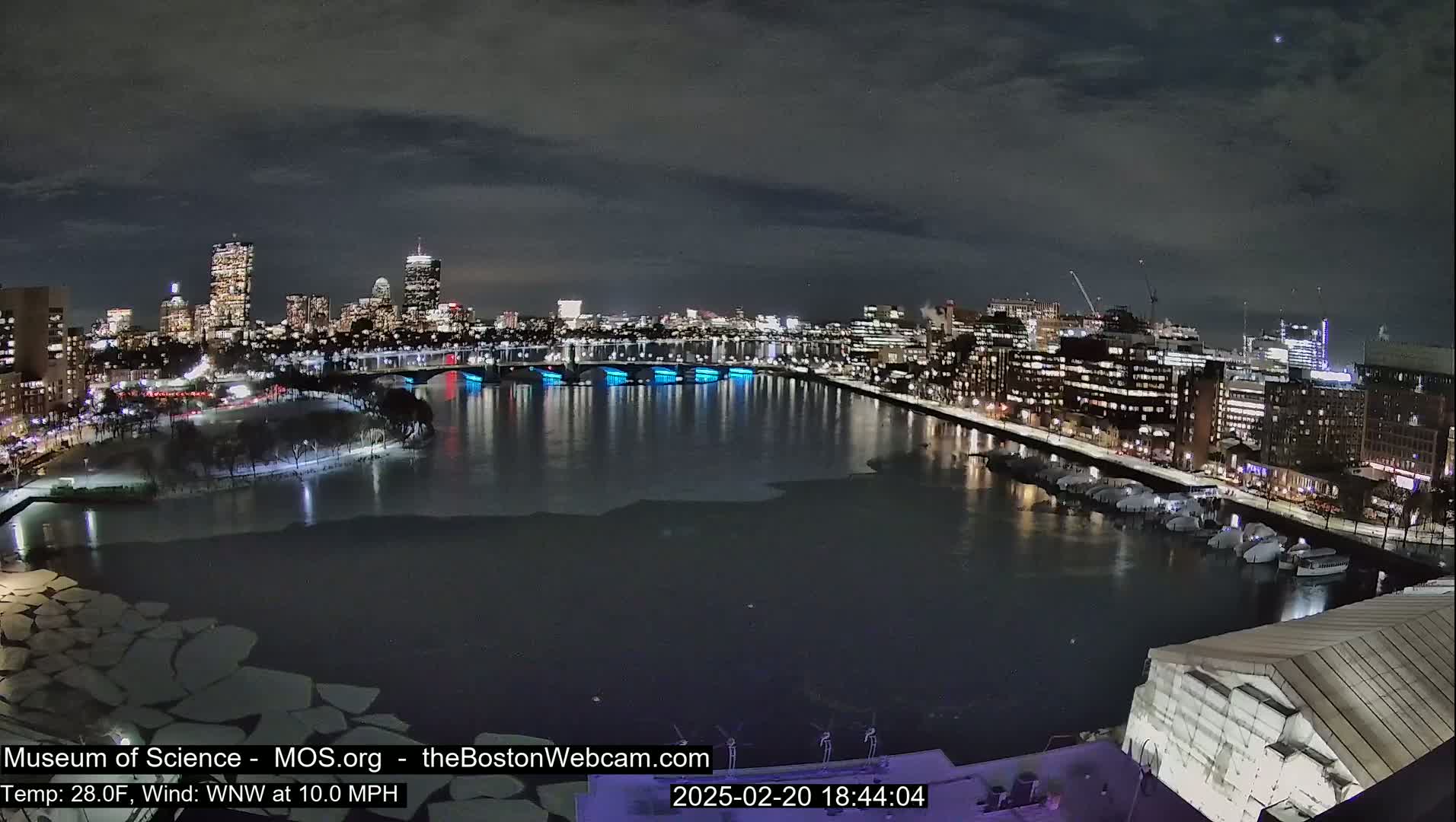 A nighttime cityscape with a partially frozen river, illuminated bridges, and a city skyline under a dark, cloudy sky.