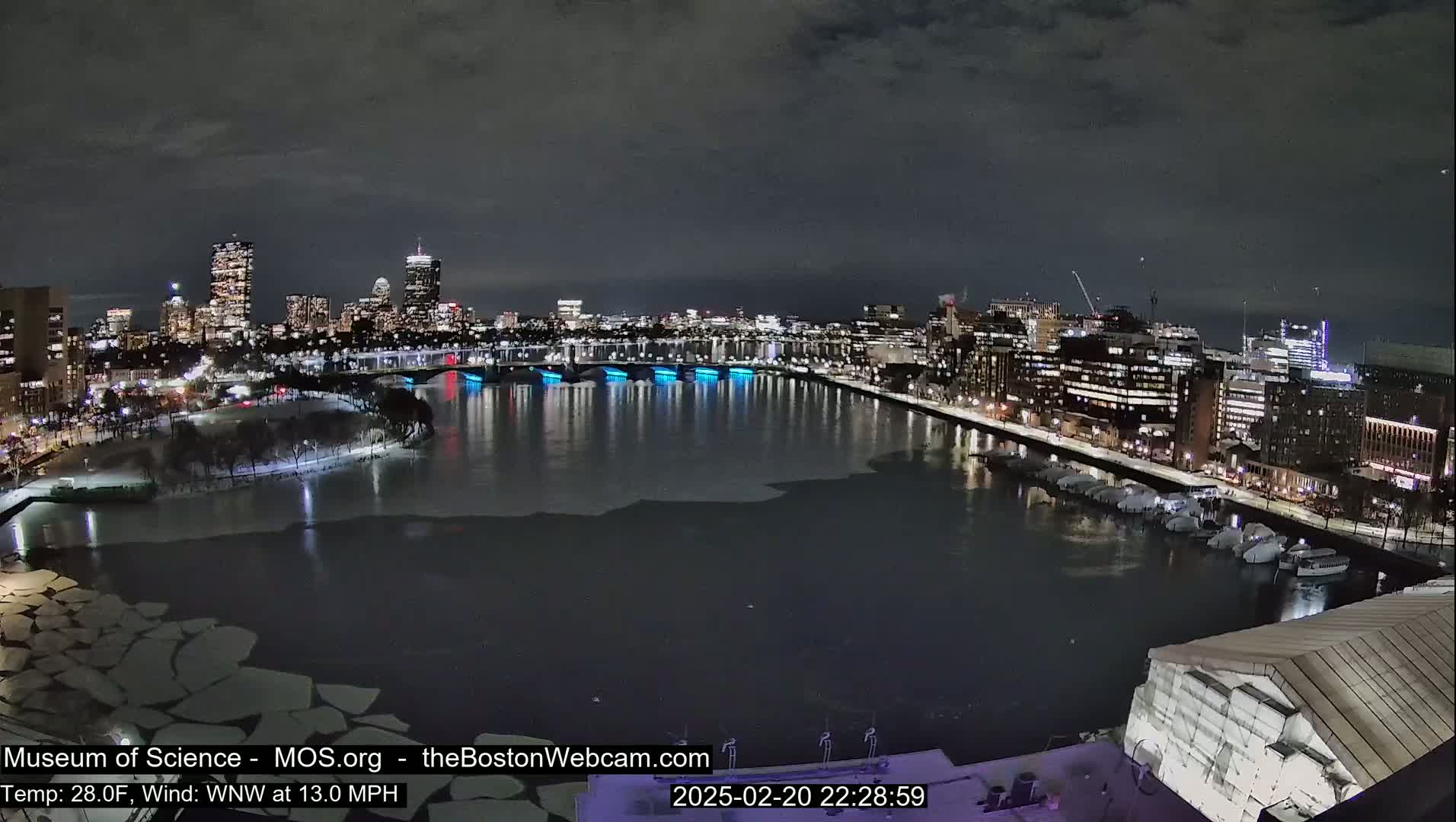 A nighttime cityscape with a partially frozen river, illuminated buildings, and a bridge with blue lights, under a dark sky.
