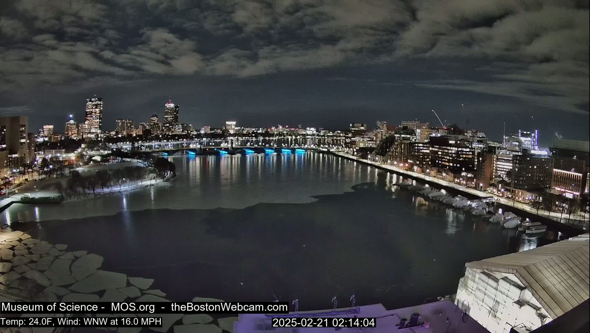 A nighttime cityscape with a partially frozen river, under a cloudy sky, and a wind of 16.0 mph.
