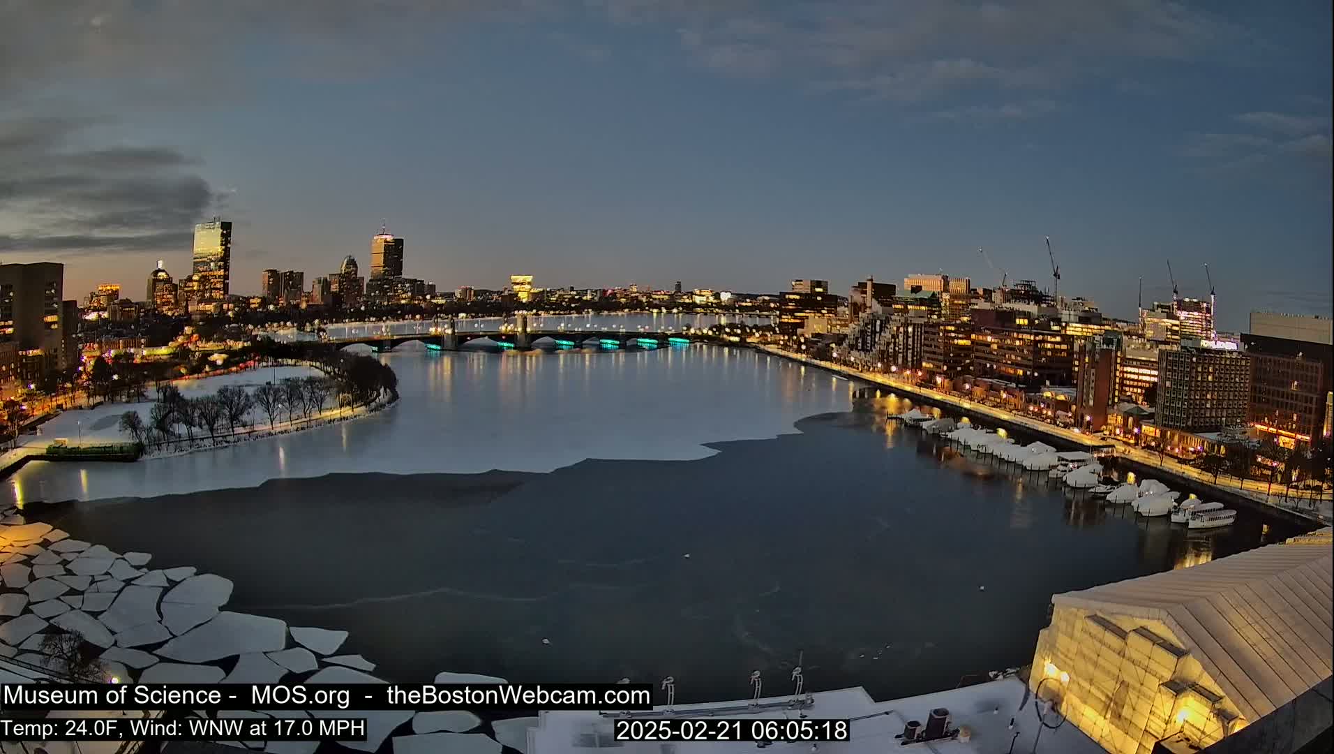 A nighttime cityscape with a river partially frozen over, showcasing city lights and a bridge under a twilight sky with a 24°F temperature and a 17 mph wind from the west-northwest.