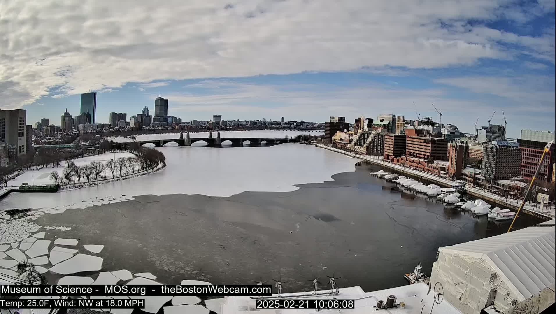 A partly frozen river flows through a snowy city, with a bridge and many buildings visible under a partly cloudy sky.