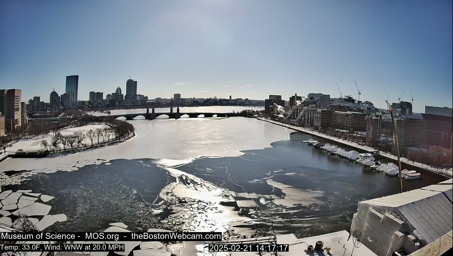 A partly frozen river flows through a city under a sunny sky, with a bridge and cityscape visible in the background.