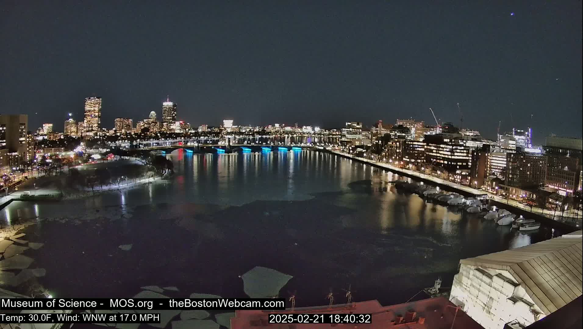 A nighttime view of a city skyline across a partially frozen river, with a moderate wind.