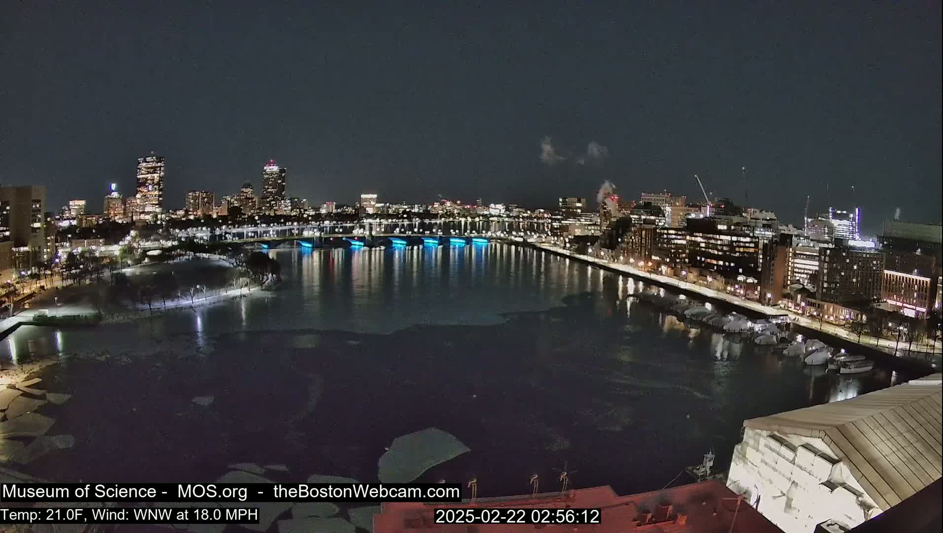 A nighttime cityscape with a river partially frozen over, illuminated buildings, and a bridge with blue lights.