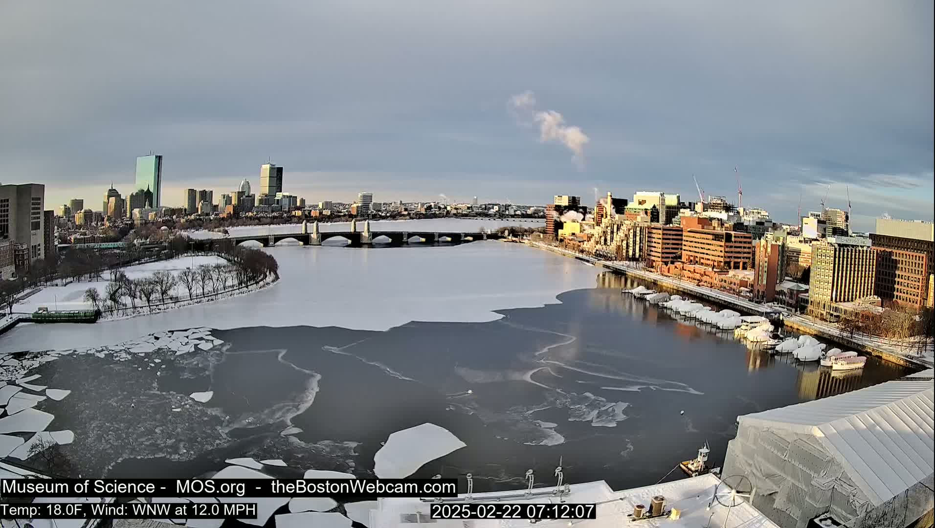 A partially frozen river flows through a city landscape under a partly cloudy sky, with snow covering the ground and some boats docked along the riverbank.
