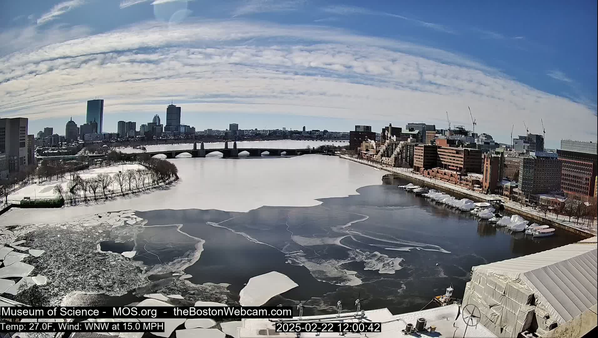 A partially frozen river flows past a city skyline under a mostly sunny sky with scattered clouds,  boats are docked along the riverbank.