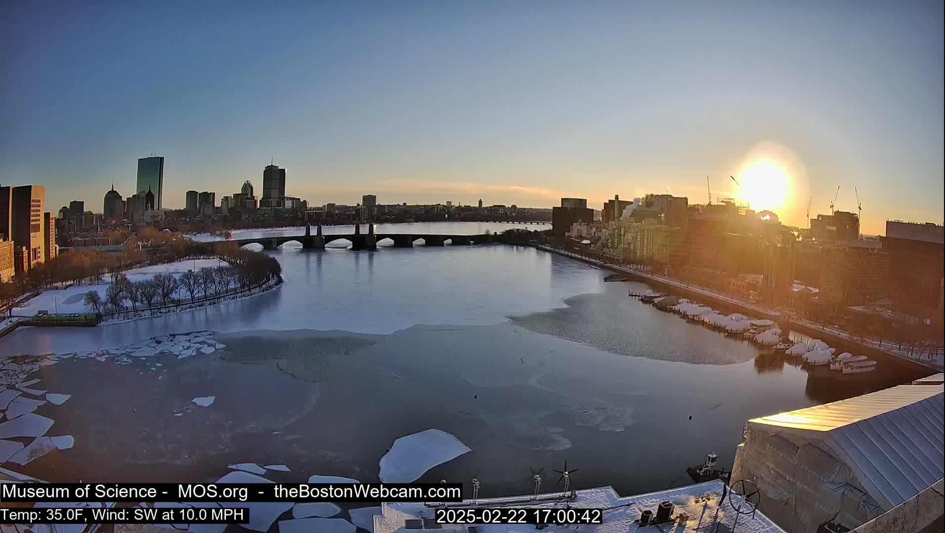 A partially frozen river flows past a city skyline at sunset, with a bridge visible in the distance and snow on the ground.