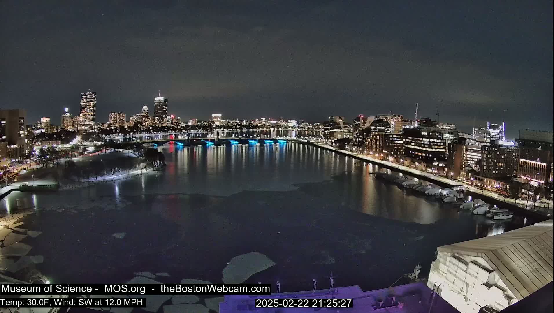 A nighttime cityscape with a partially frozen river, illuminated bridges, and a city skyline under a clear sky, with a southwest wind at 12 mph and a temperature of 30°F.