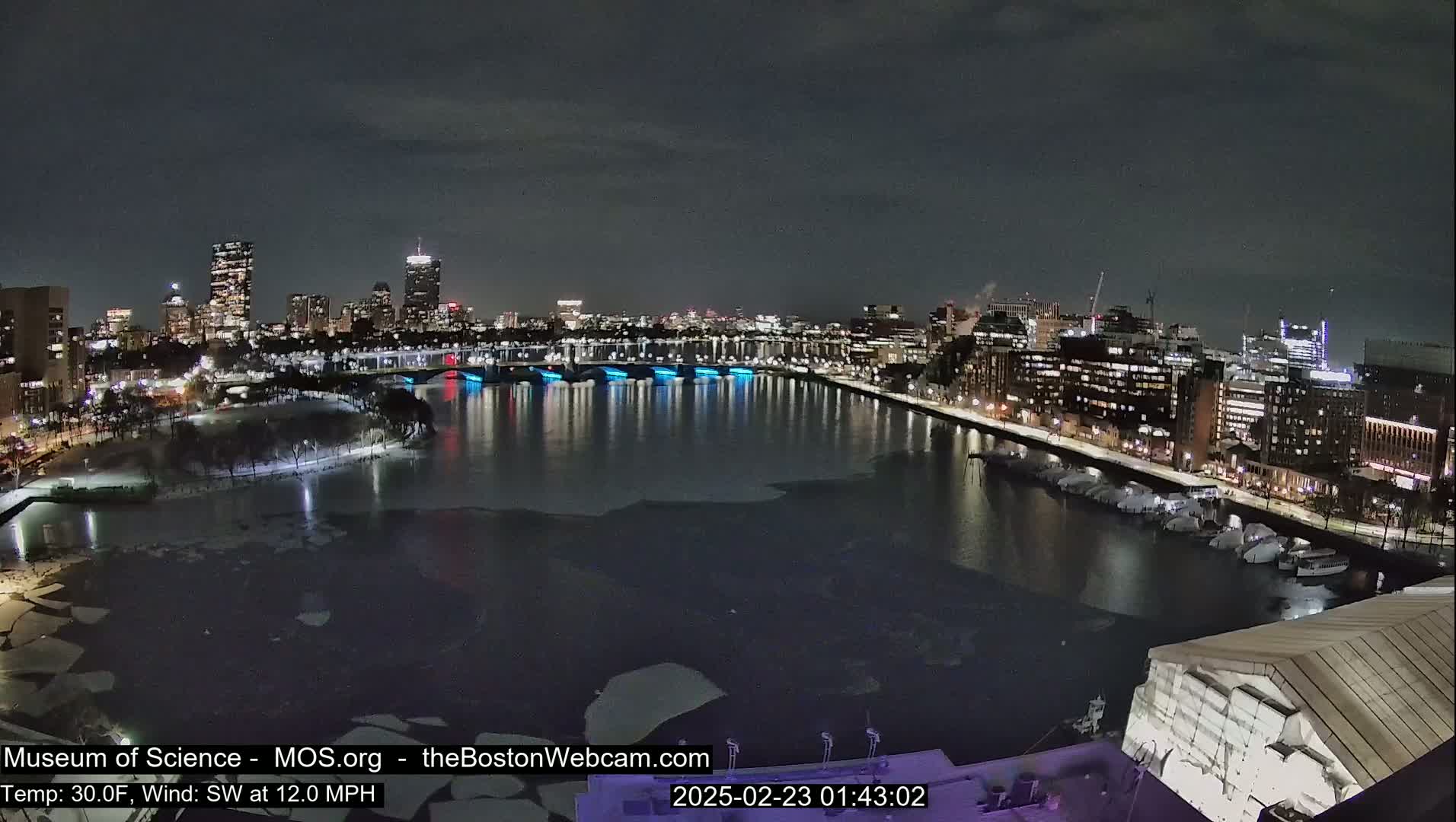 A nighttime view of a city skyline across a partially frozen river with a southwest wind at 12 mph and a temperature of 30°F.